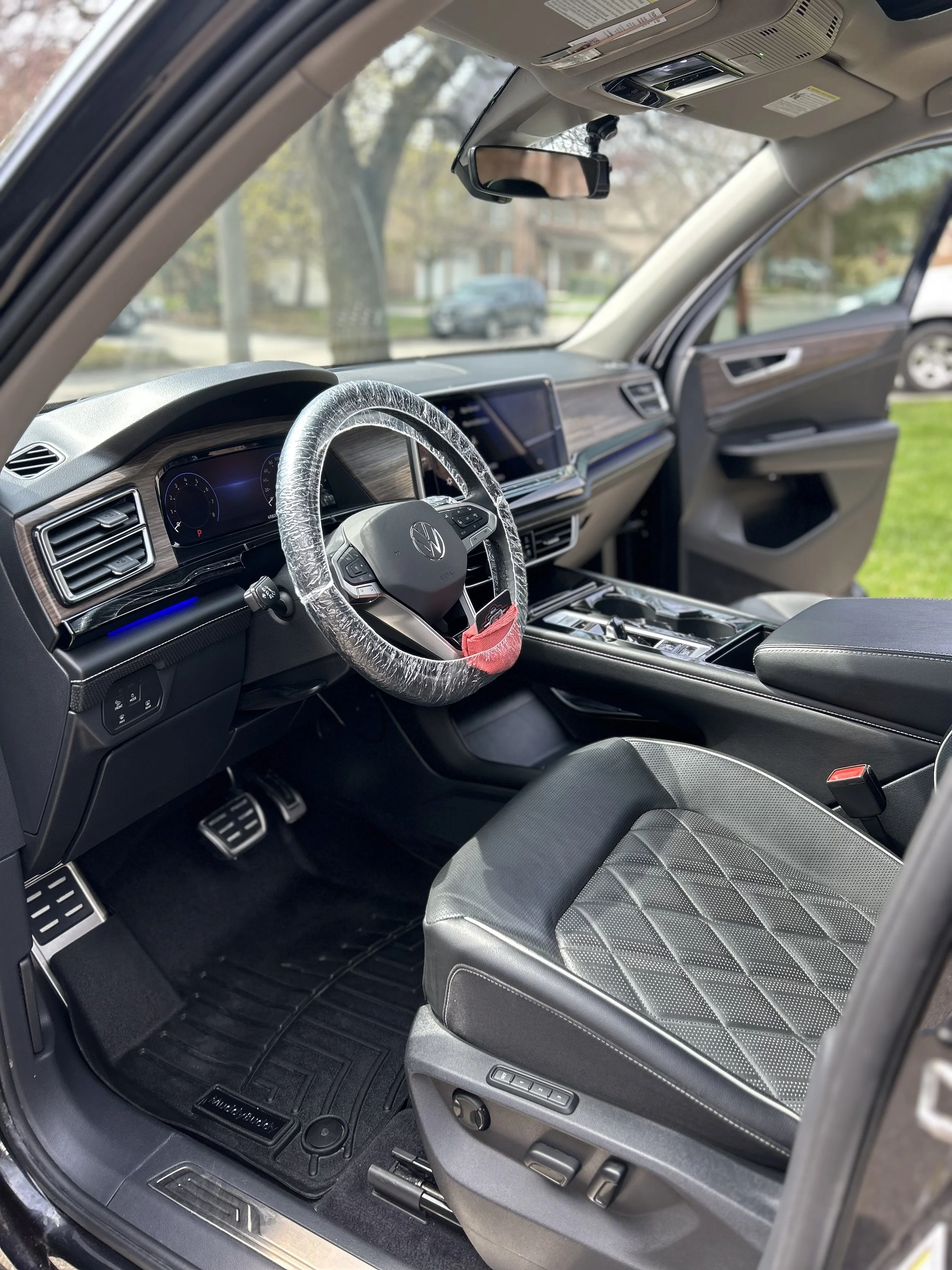 The interior of a modern vehicle, showing the driver's seat, steering wheel with protective plastic cover, dashboard, gear shift, and front passenger seat. The dashboard features digital displays and controls, and the car is parked outside with trees