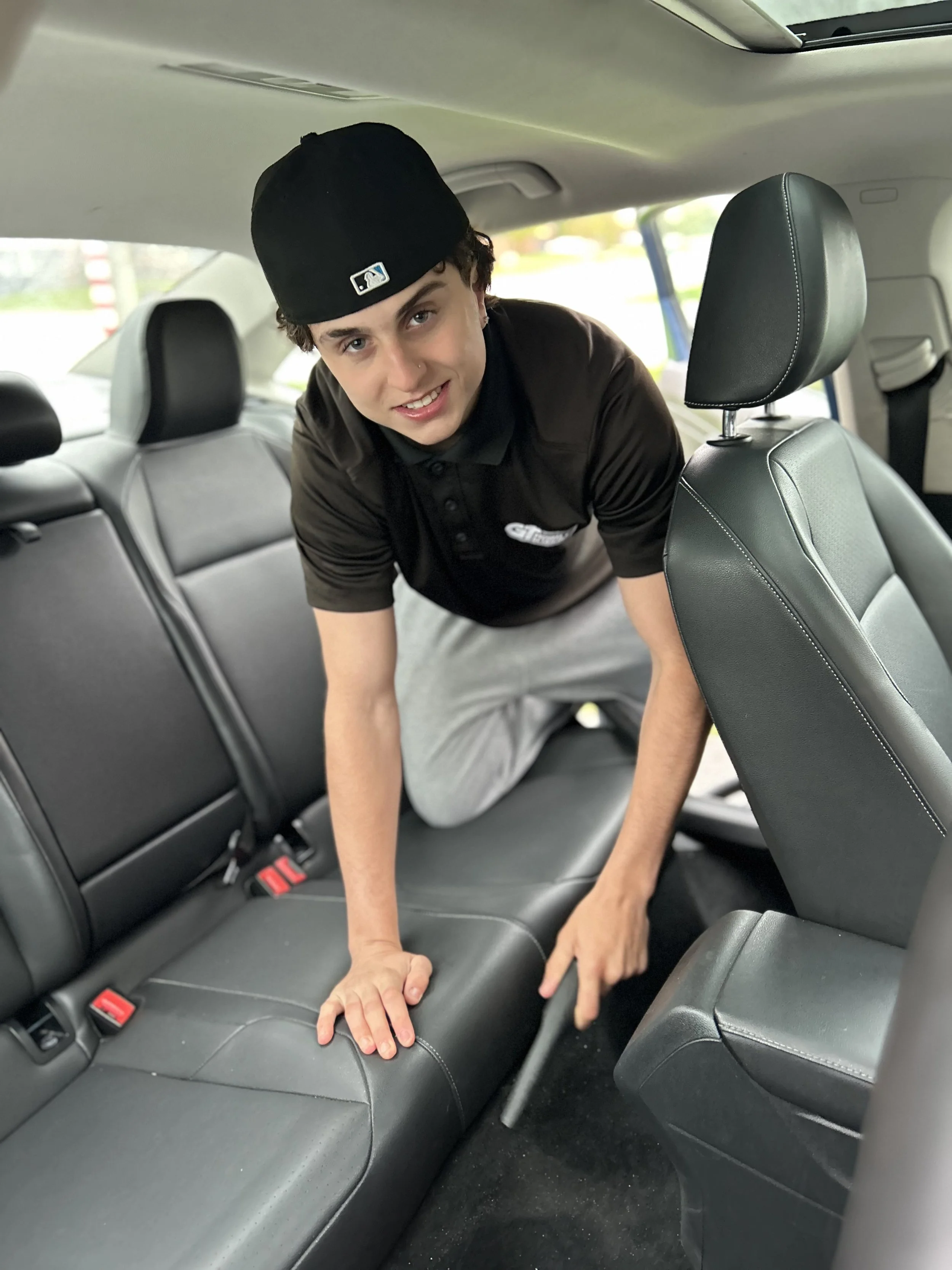Young man kneeling in the back seat of a car, adjusting the collapsible car seat in a black shirt and a black baseball cap.