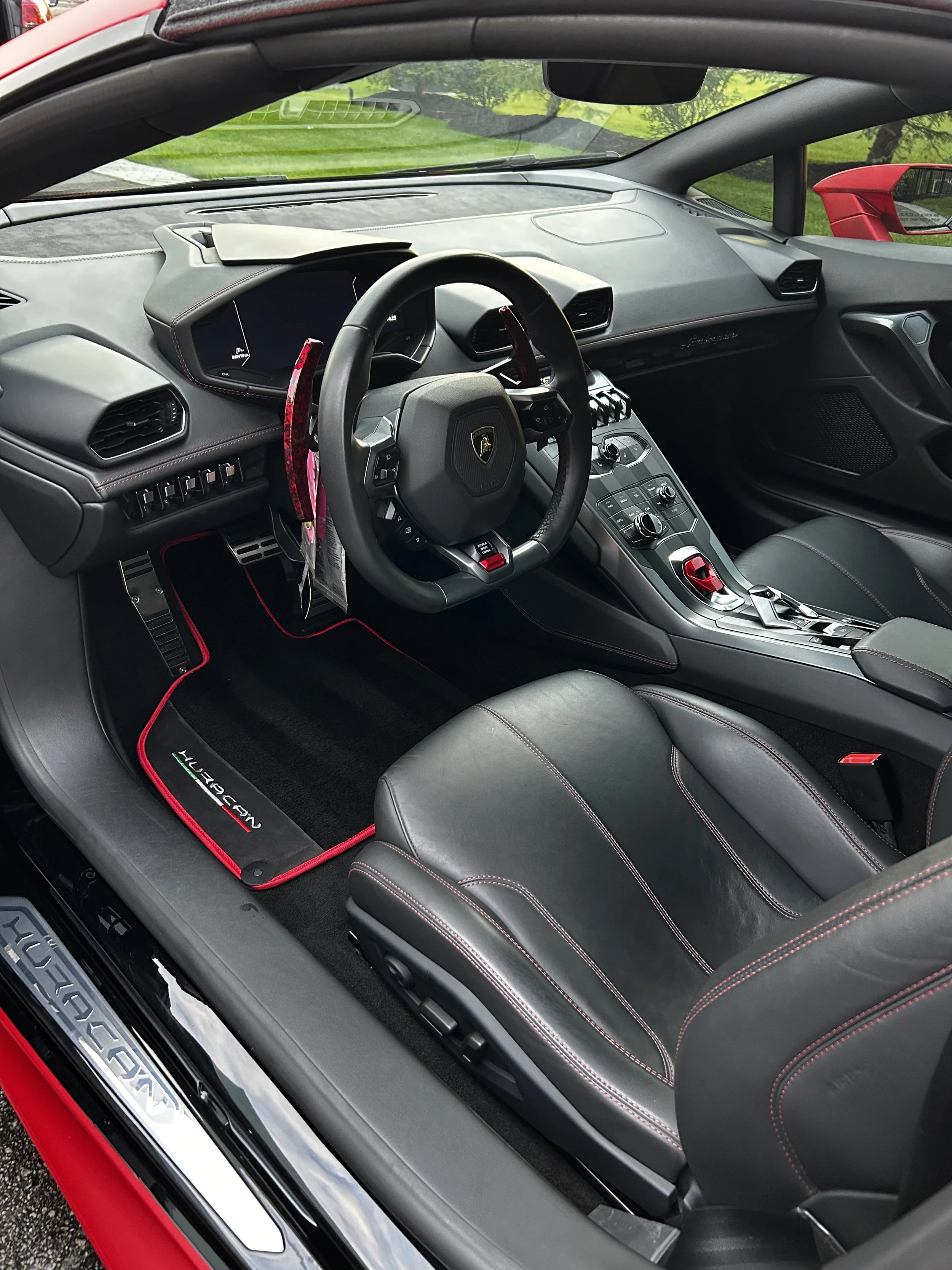 Interior view of a Lamborghini Huracán with black leather seats, red stitching, a steering wheel with Lamborghini emblem, and a red start button, with a black and red floor mat labeled 'Huracán'.
