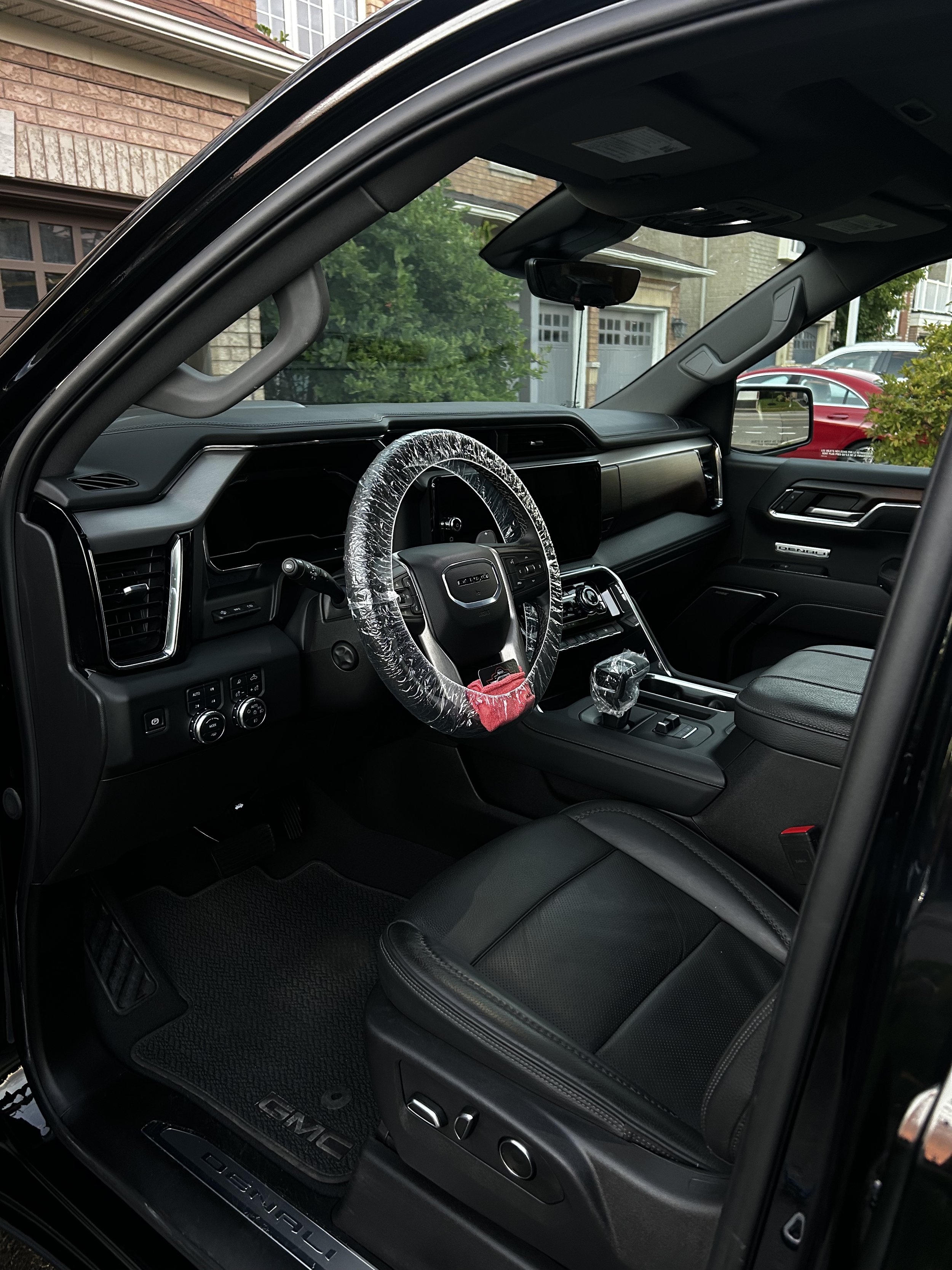 Interior of a black GMC vehicle with a plastic-wrapped steering wheel and black leather seats, parked in a driveway with residential buildings and greenery outside.