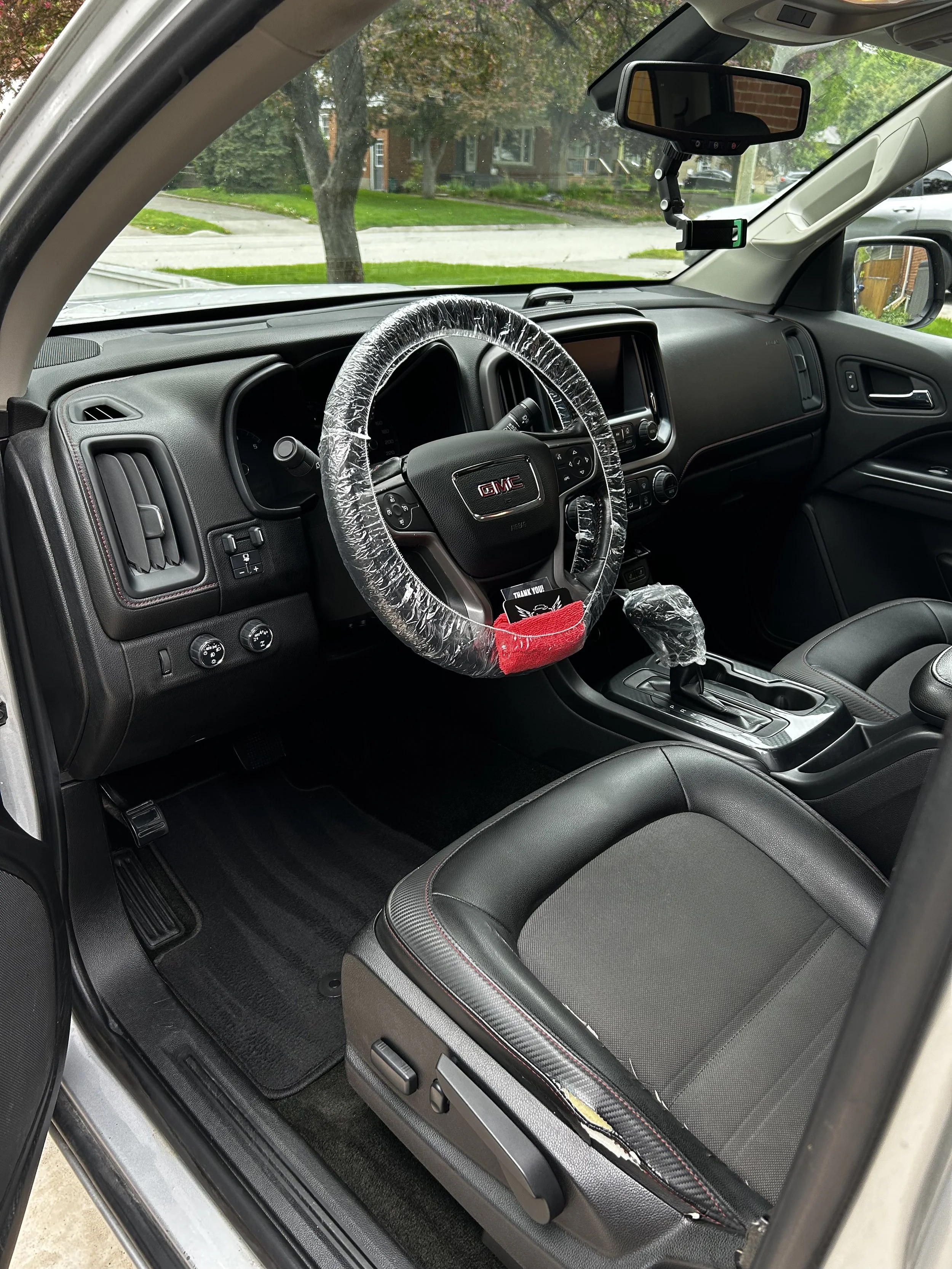 The interior of a GMC vehicle showing the dashboard, steering wheel with a plastic cover, gear shift, and front passenger seat. The outside view through the windshield shows a residential neighborhood with trees and houses.