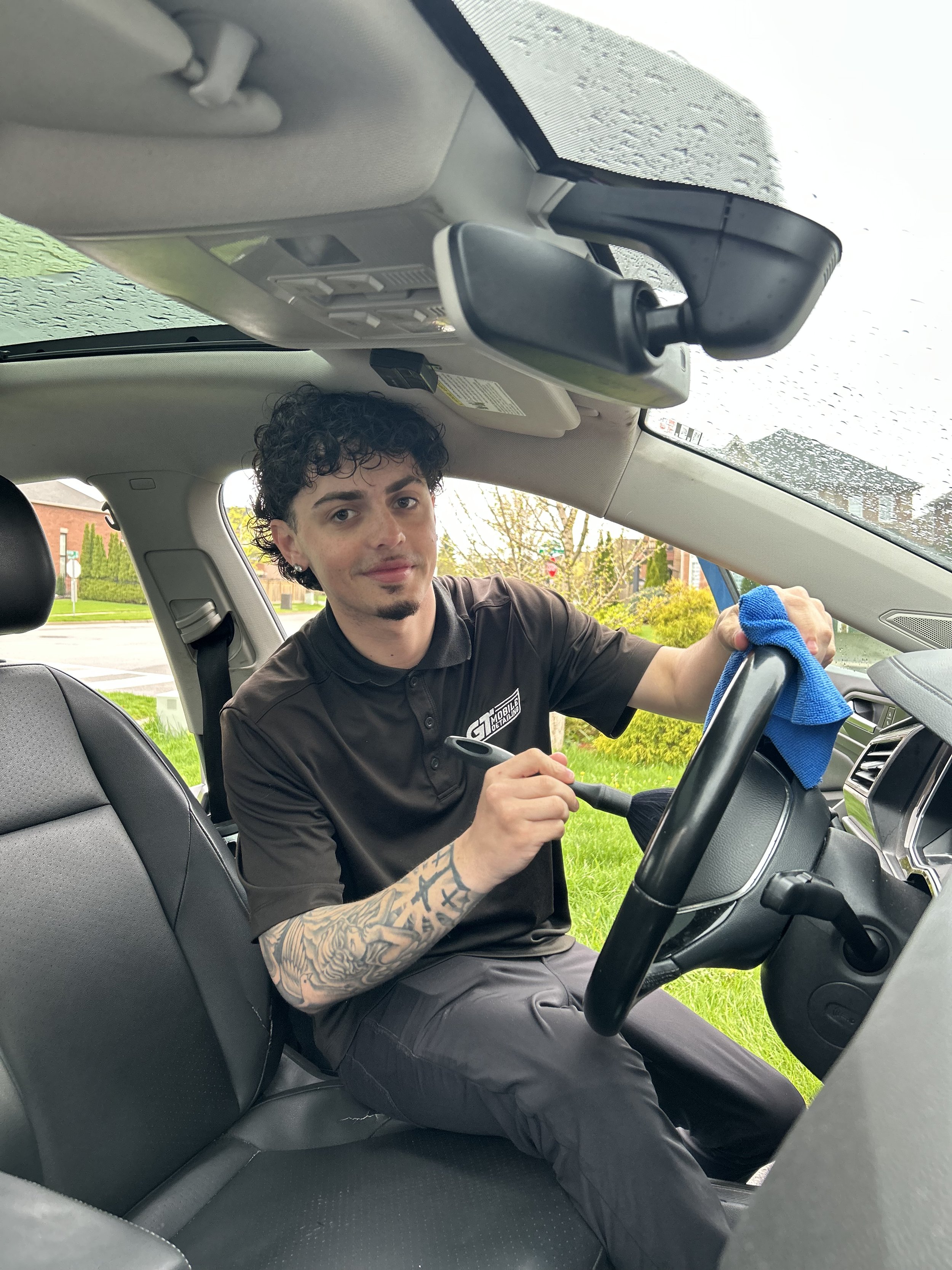A young man with curly dark hair and tattoos on his right arm is cleaning the interior of a car with a blue cloth. The image is taken from the backseat, showing the front passenger seat and the man sitting in the driver's seat, with a street scene visible through the windshield.