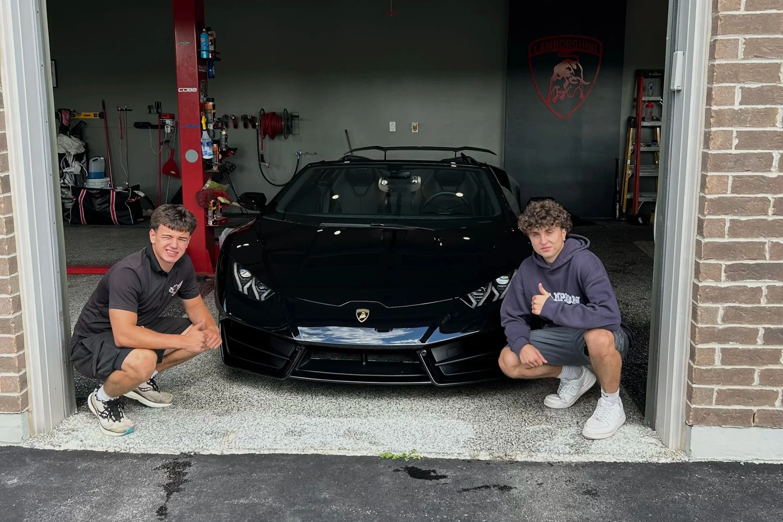 Two young men squatting in front of a sleek black Lamborghini sports car inside a garage. One is wearing a black polo shirt and shorts, and the other is in a gray hoodie and shorts, giving thumbs up. The garage has tools, a ladder, and a Lamborghini logo on the wall.