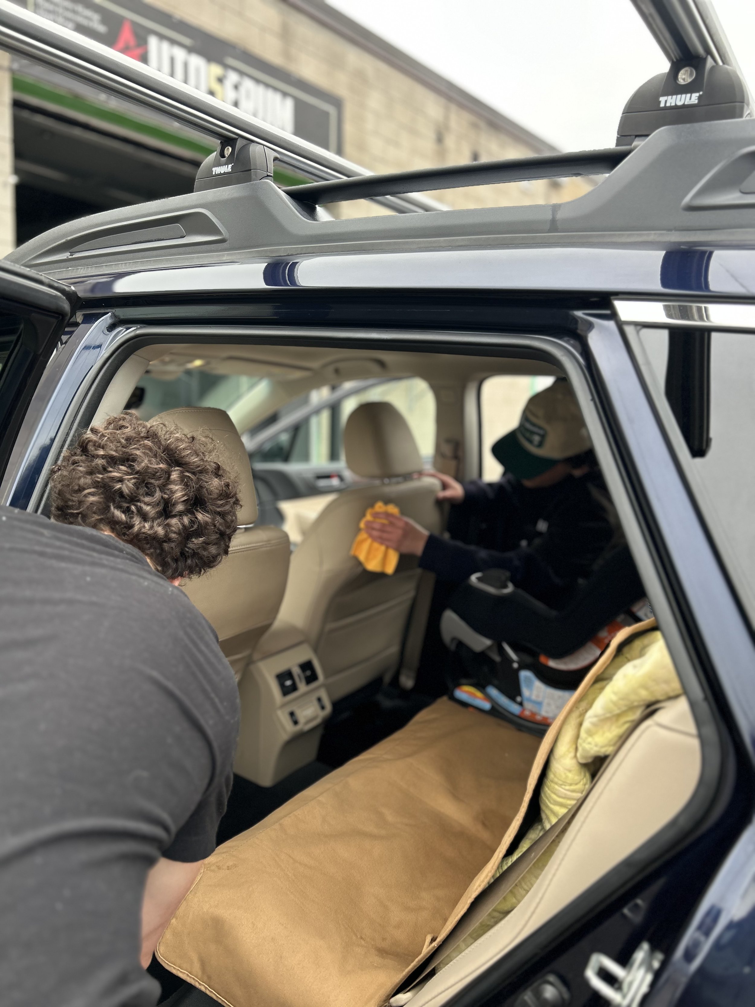 Inside a vehicle, a person is placing a beige cushioning pad on the back seat. Two children are seated, one in the front and one in the back, with the child in the back wearing a cap and holding a small object. The vehicle's interior shows beige seats and rear air vents, with an open door revealing a brick building and a vehicle outside.