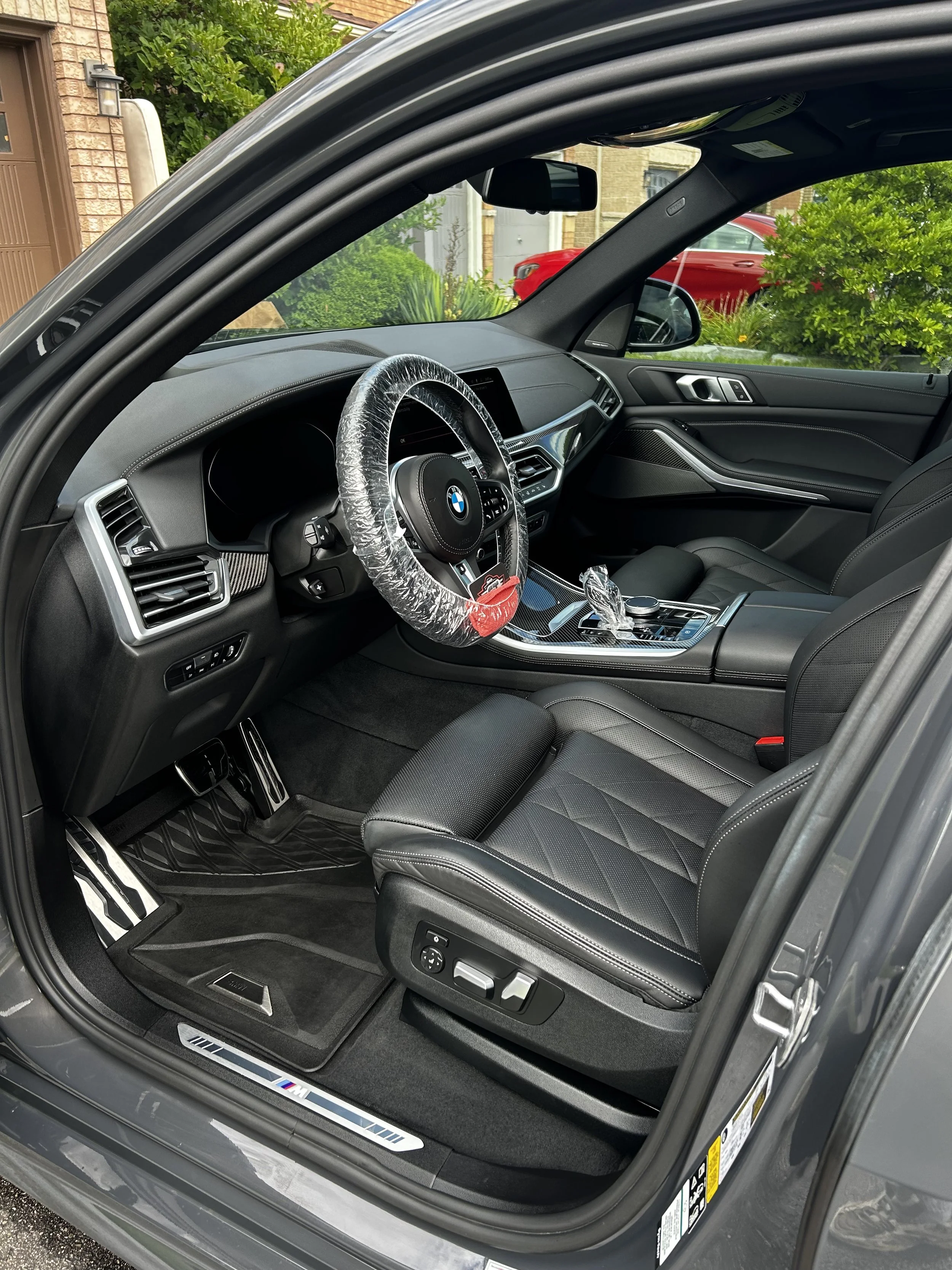 The interior of a modern black BMW vehicle, showing an unlocked door, black leather seats, dashboard, steering wheel with a plastic cover, and a gear shift, with a view of a driveway and garden outside.