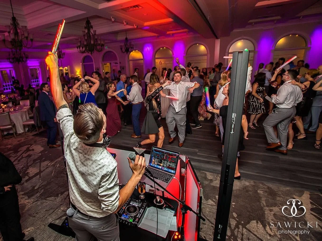 People dancing at a party or wedding reception in a large, elegant room with colorful lighting and a DJ playing music at the front.