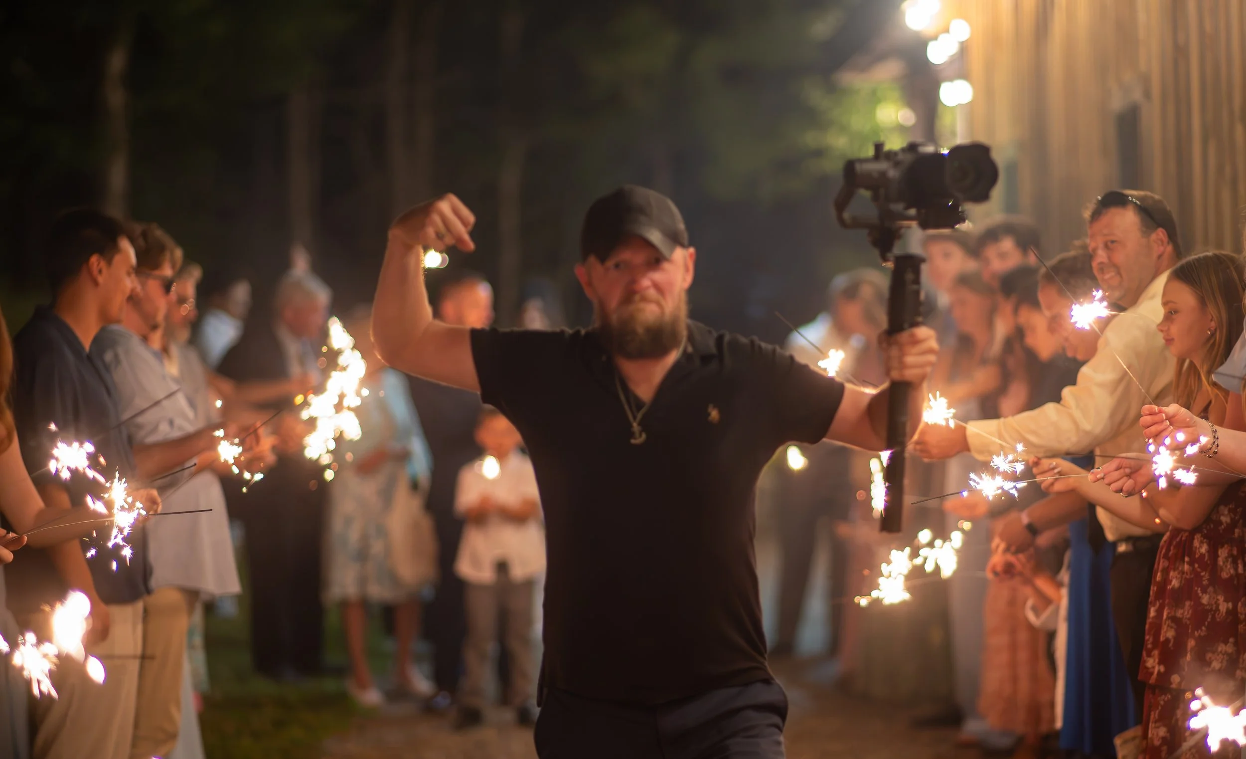 A group of people celebrating at night with sparklers, standing in a line outdoors with trees and lights in the background. A man in the center flexes his arm and smiles.