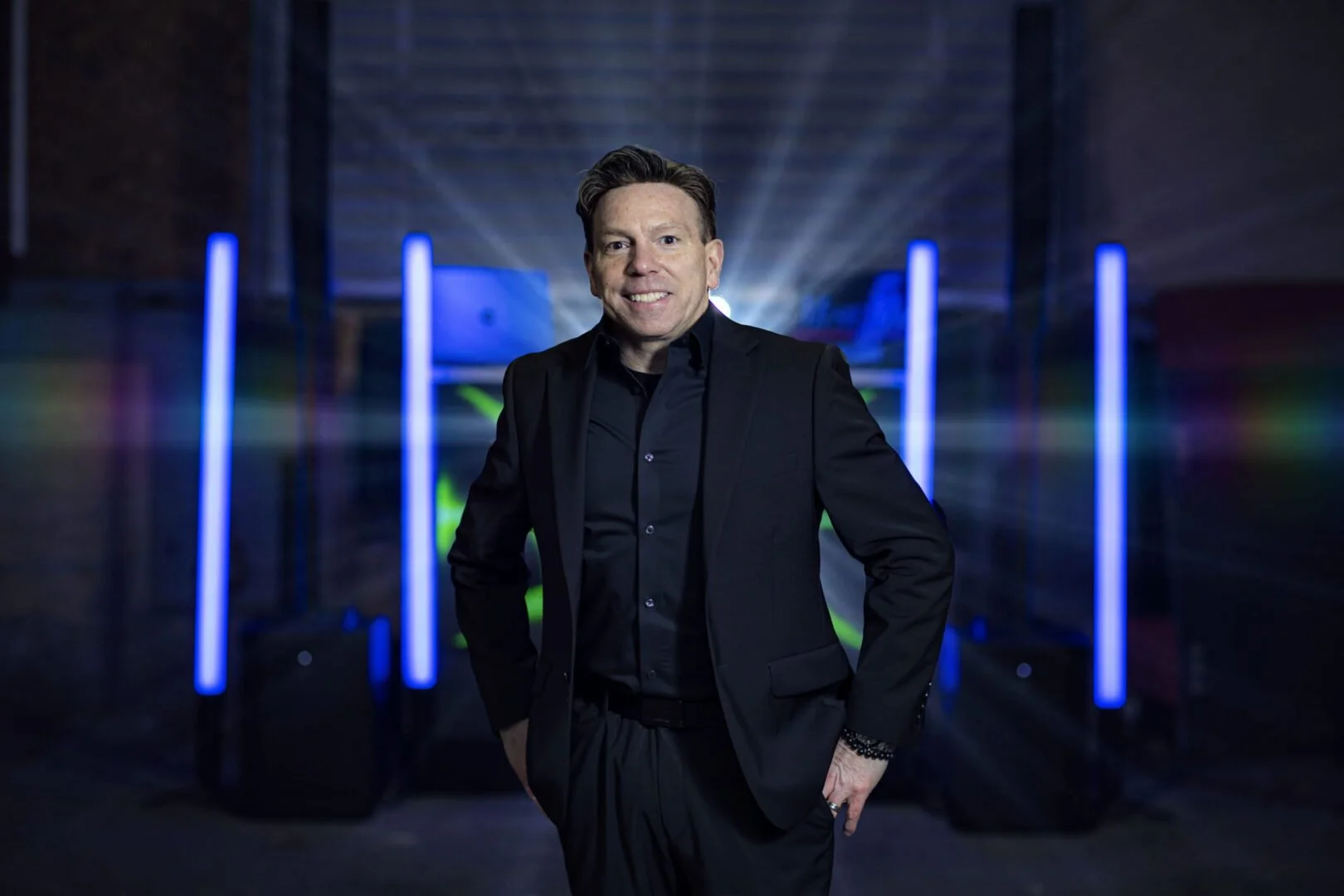 A smiling man in a black suit standing in front of a futuristic industrial or technological background with blue lights and laser beams.