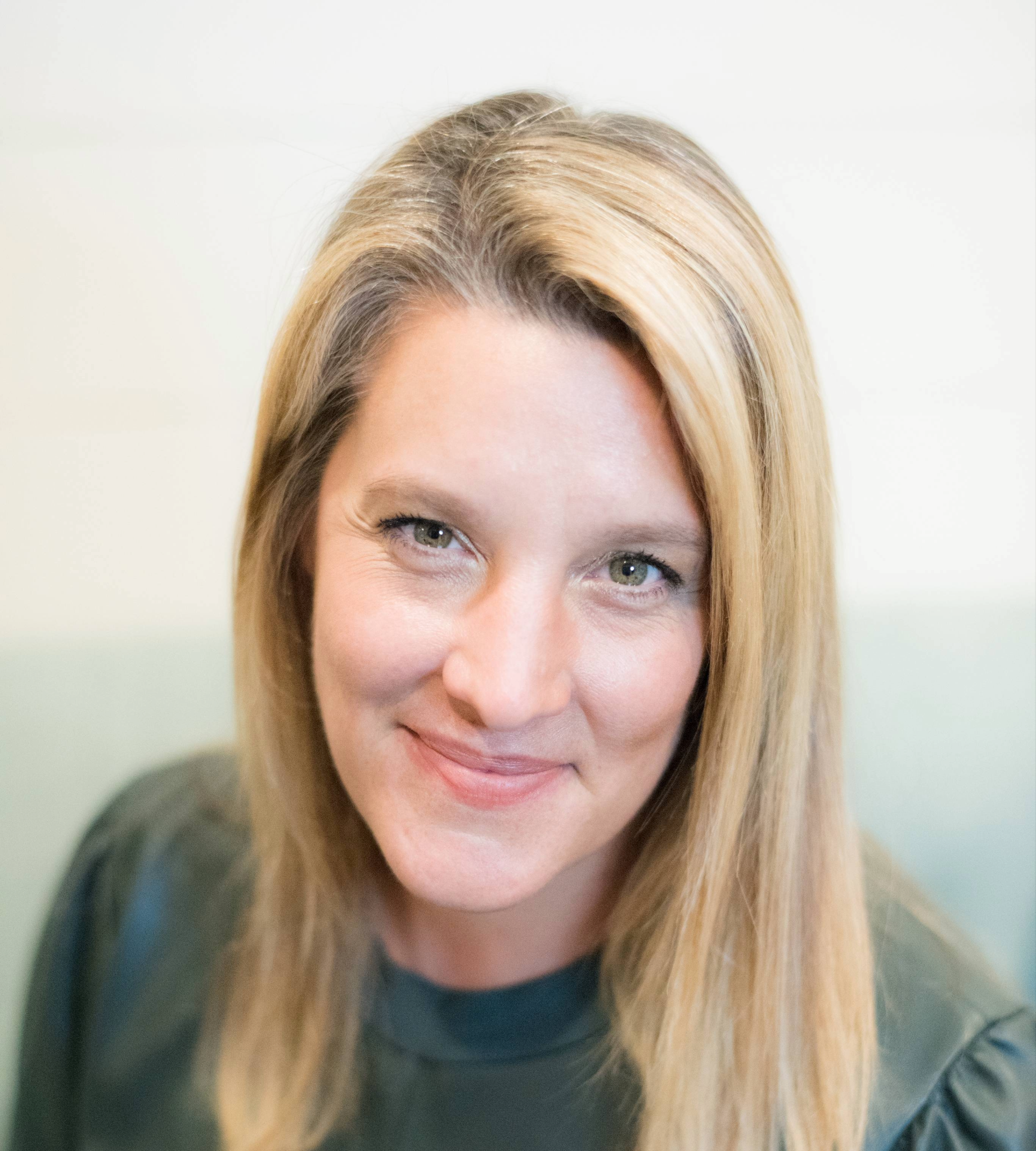 A woman (Abbey Danczak) with long blonde hair and light skin smiling at the camera, wearing a dark top.