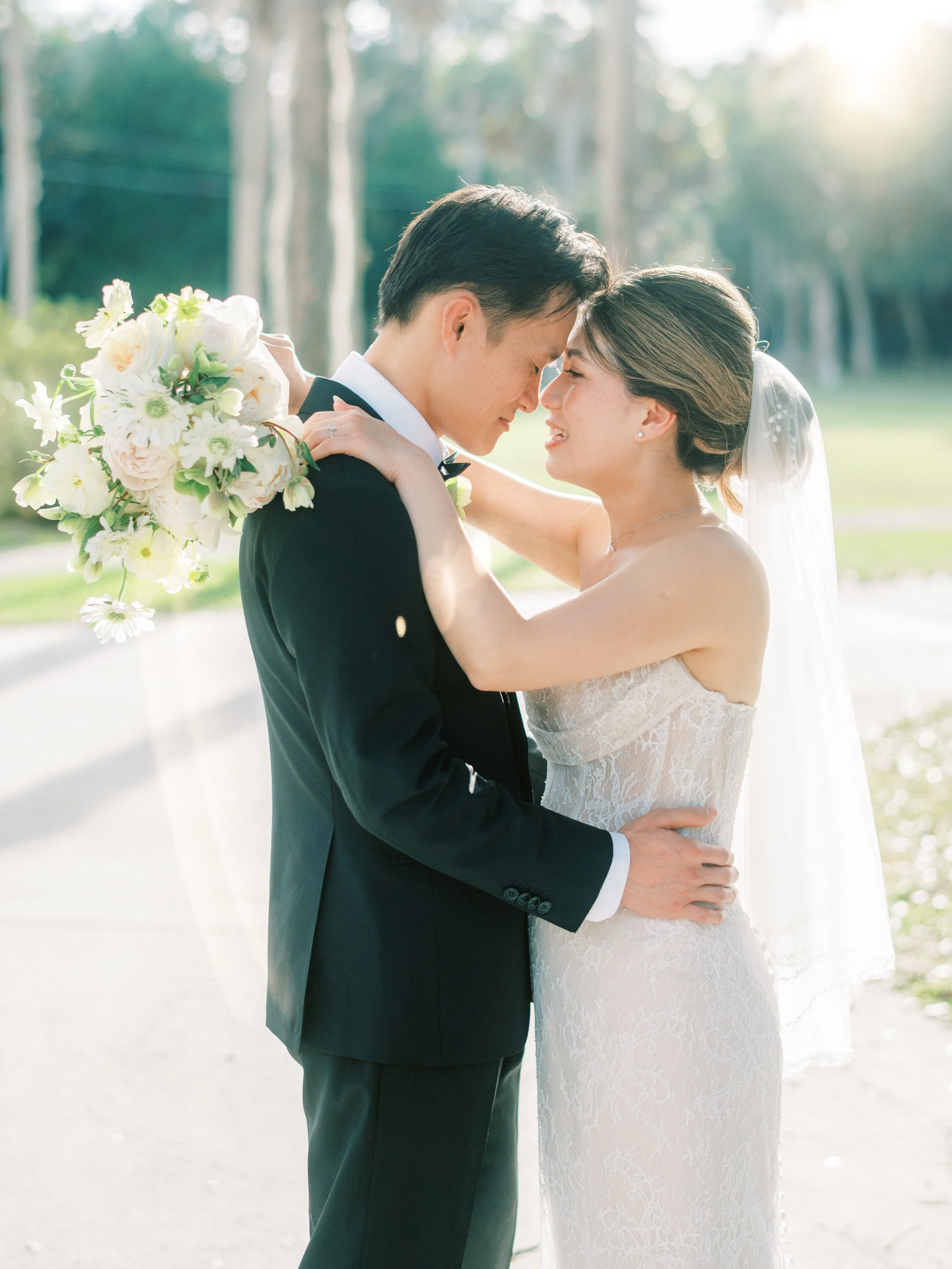 A man and a woman are jumping into a swimming pool at night, holding hands. The woman is wearing a sleeveless dress, and the man is wearing a dress shirt and pants. There are people in the background near a building and bar area, with some watching and taking pictures.