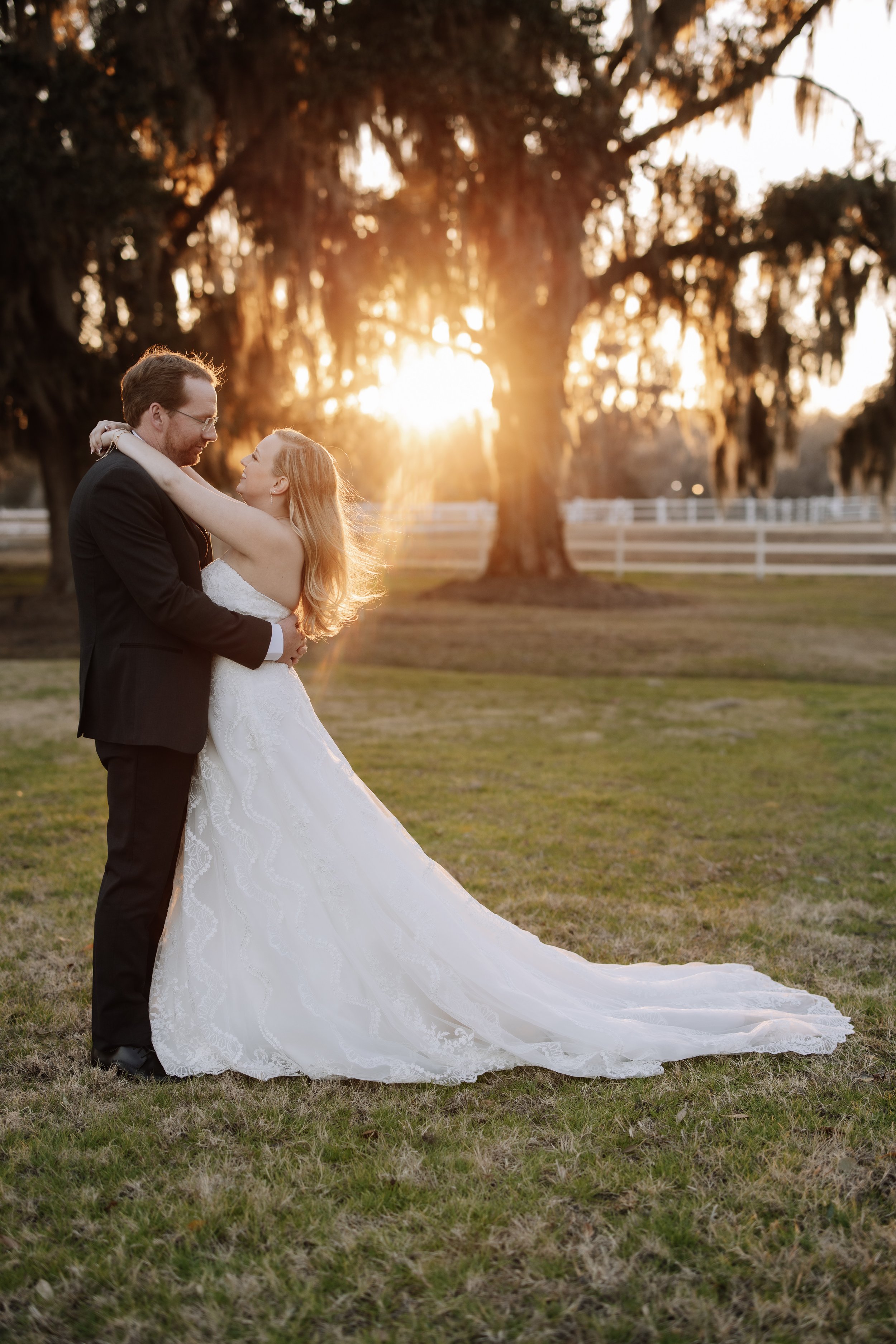 A black and white photo of a wedding couple outdoors in a garden. The bride, in a wedding dress and veil, is kissing the groom, who is seated on a bench in a light suit. The background has trees and bushes.