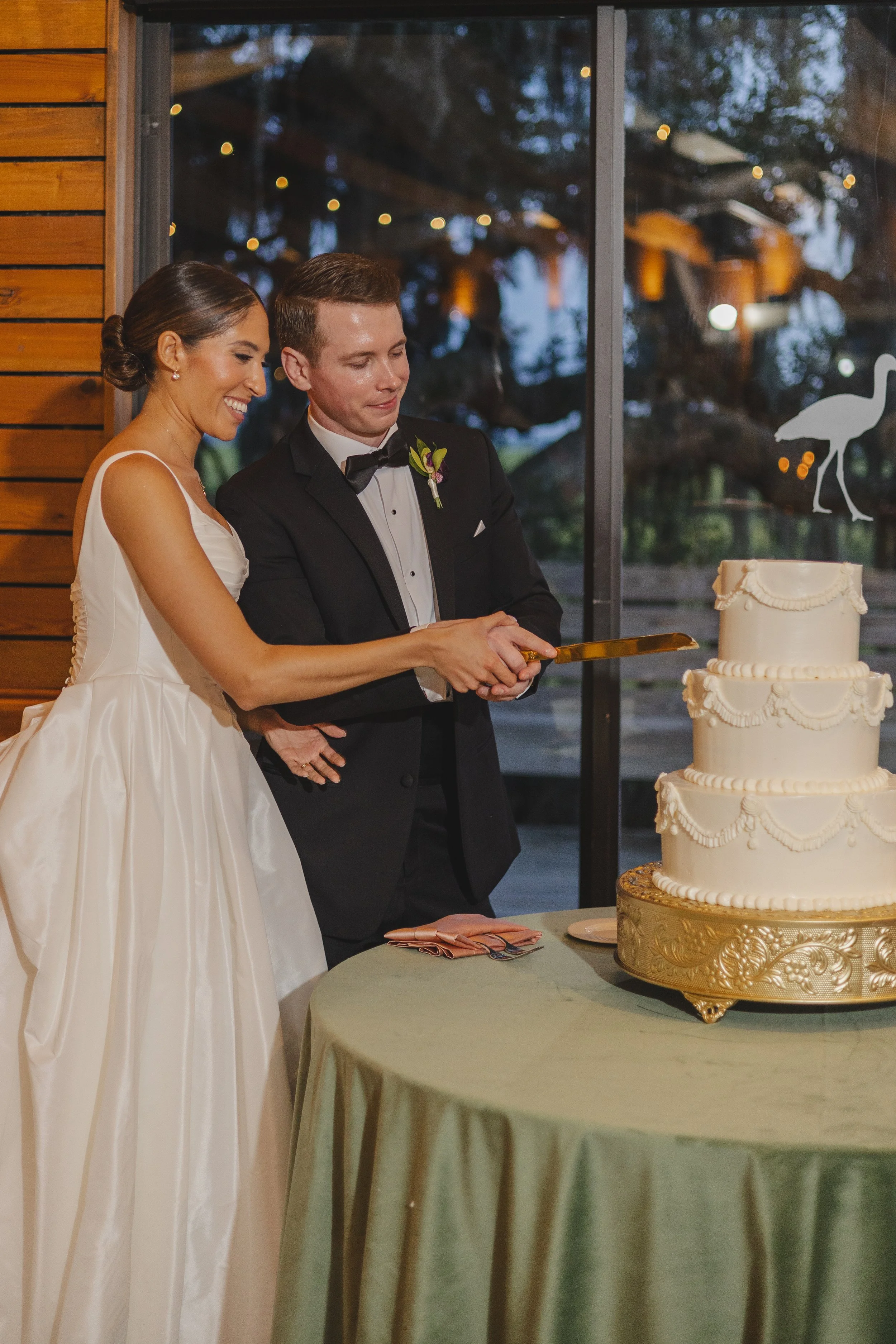 Happy newlywed couple holding hands after wedding ceremony outdoors in a wooded area, the bride in a strapless white wedding dress holding a colorful bouquet, and the groom in a beige suit with brown shoes, surrounded by friends and family.