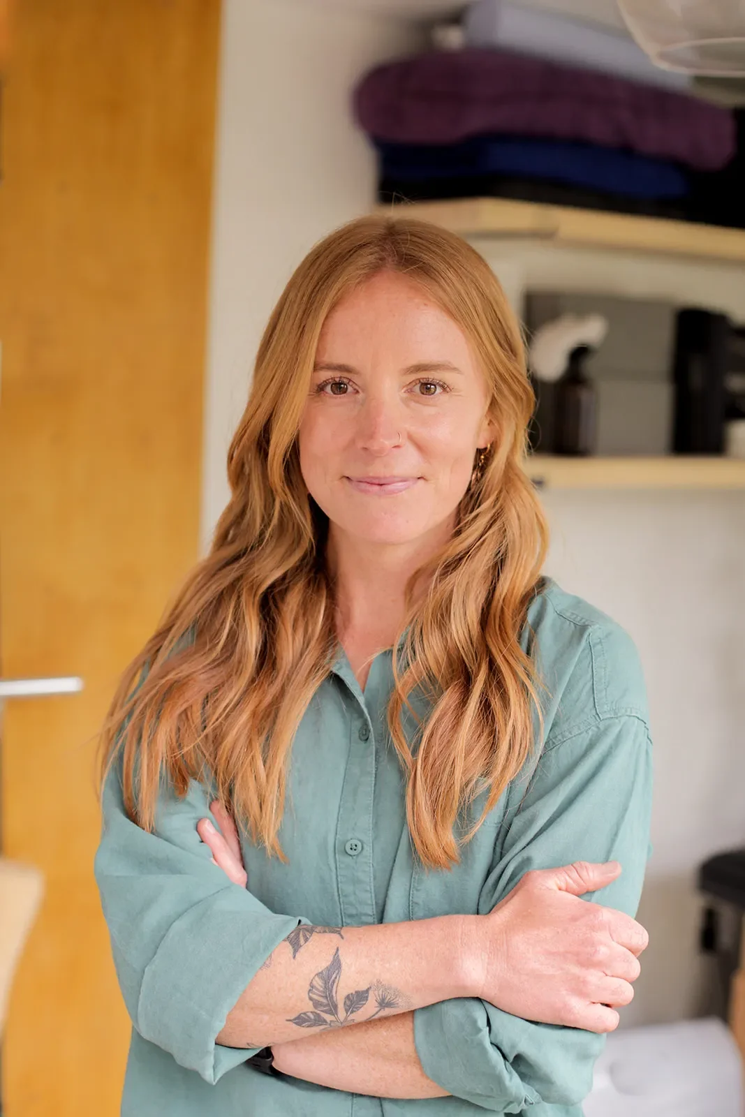 A woman with long red hair, wearing a green shirt, is standing with arms crossed in a room with folded clothes on a shelf behind her.