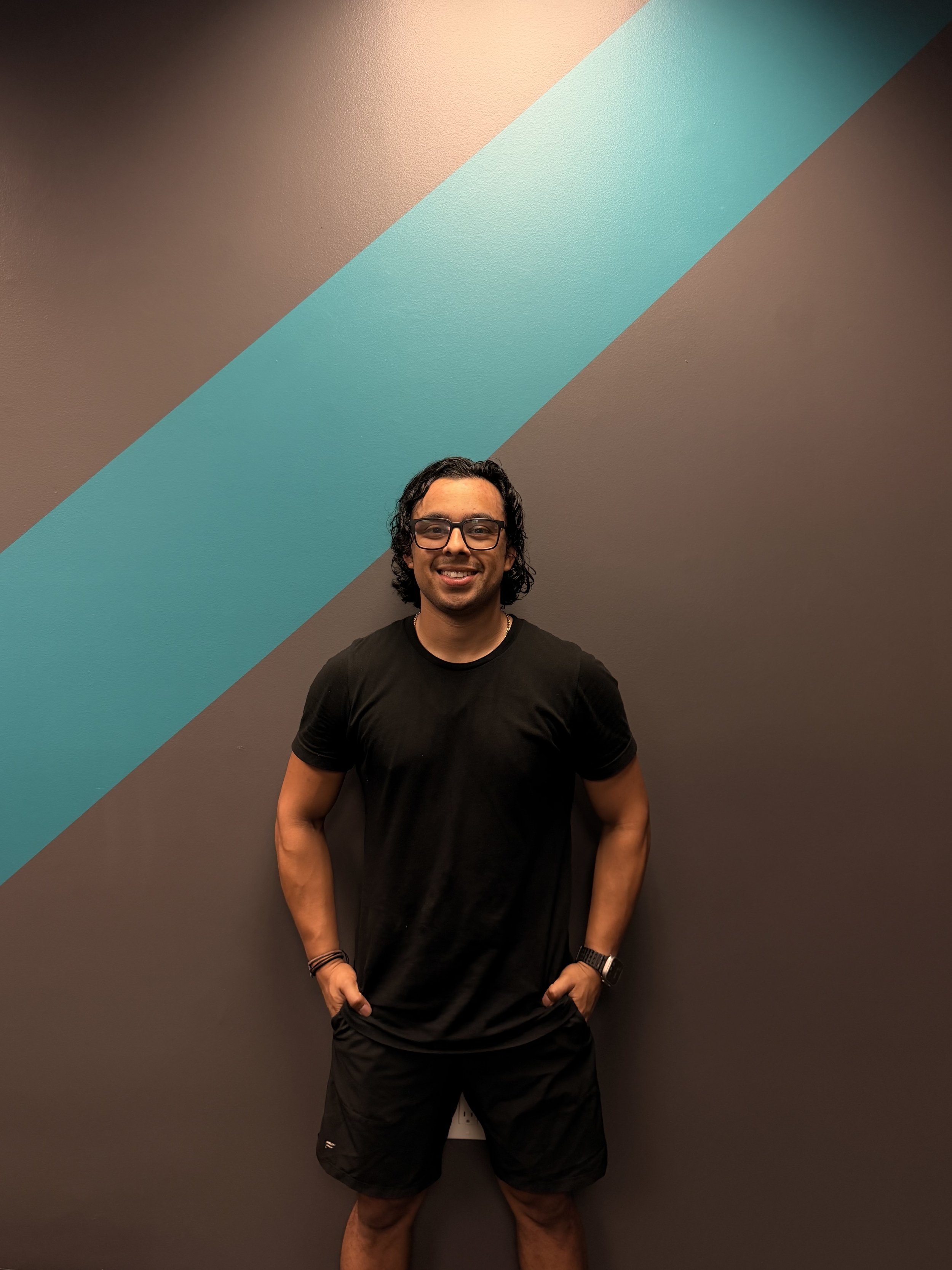 A man with glasses and long curly hair standing in front of a wall with diagonal colorful stripes, wearing a black t-shirt and black shorts.