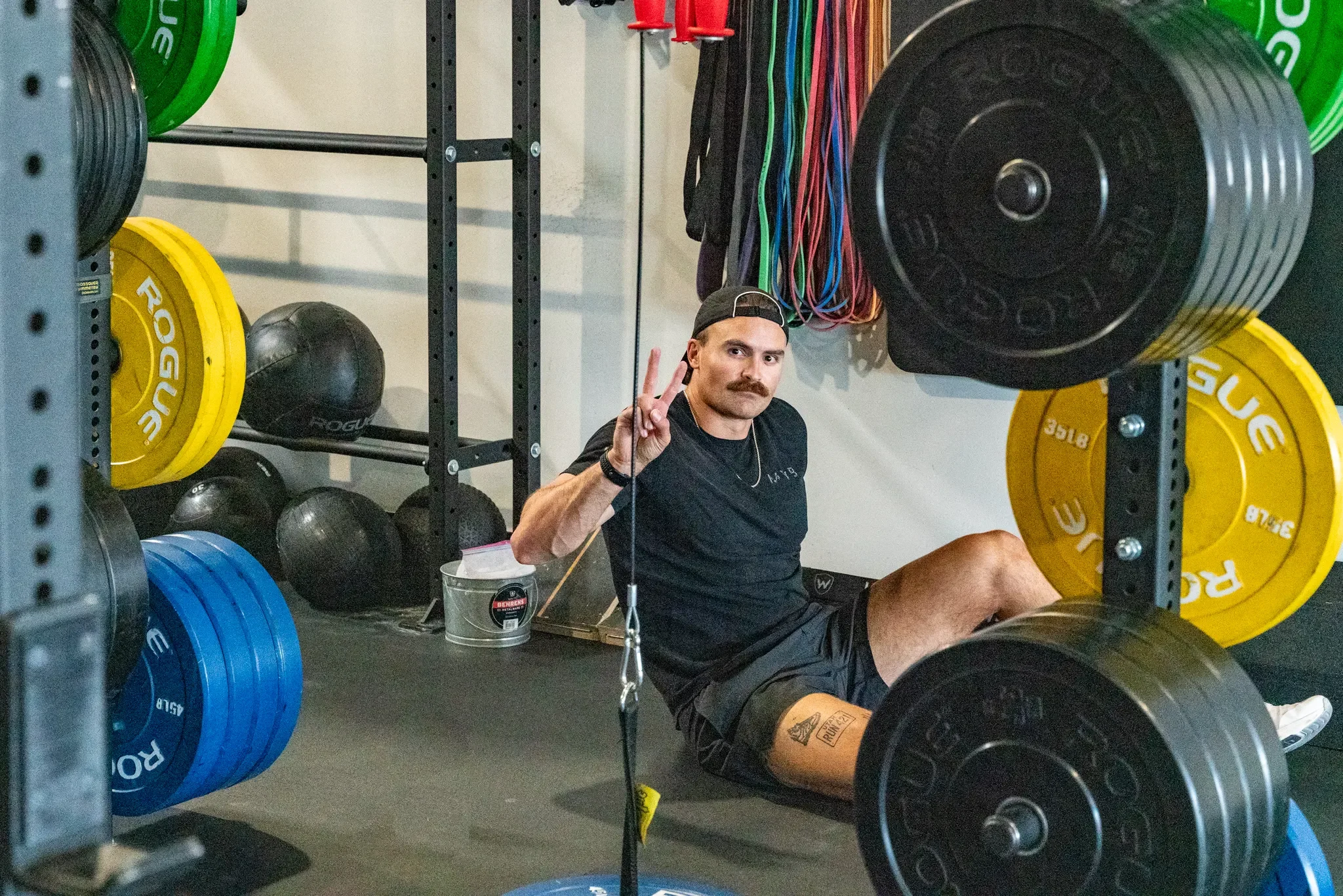 A man with a mustache and a backwards cap sitting on the floor of a gym, making a peace sign with his right hand, surrounded by weightlifting equipment and colorful resistance bands hanging on the wall.