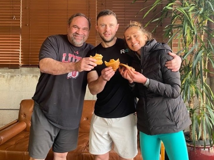 Three people standing closely together, smiling, and holding sandwiches in a cozy indoor setting with a brown leather couch, wooden window blinds, and a tall green plant.