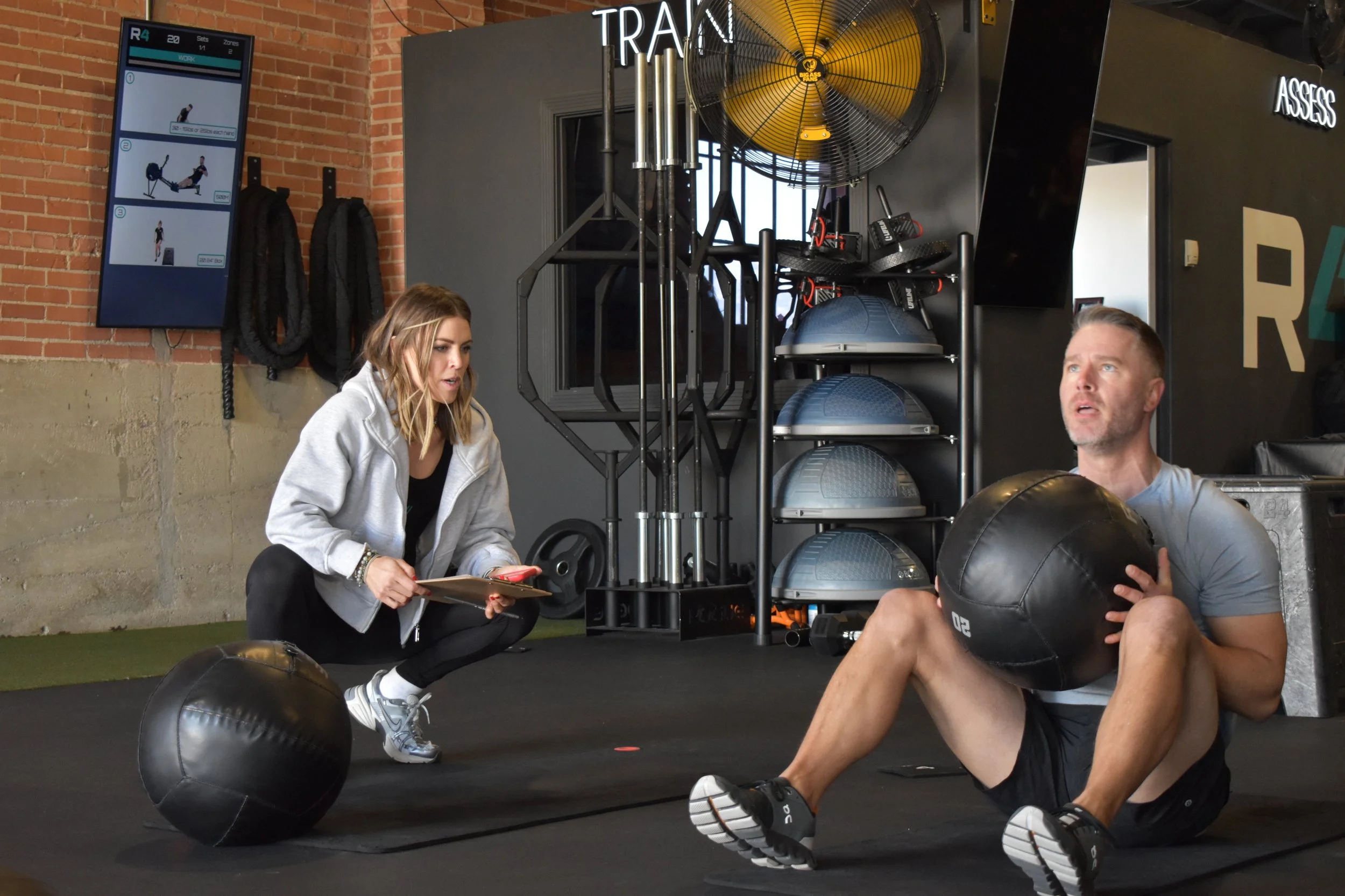 A man and woman working out in a gym studio. The man is sitting on the floor holding a black medicine ball, and the woman is crouching nearby, holding a clipboard and pen, coaching him. Exercise equipment and a wall with a digital display are visible in the background.