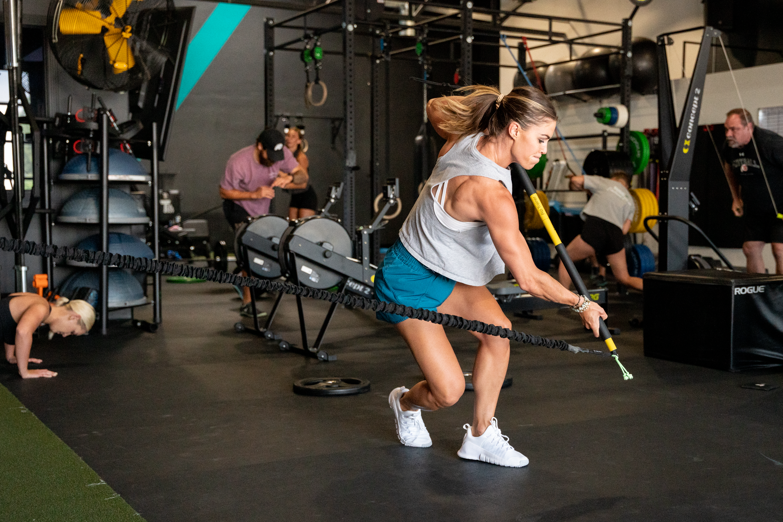 Woman doing sled pulls in a gym with resistance bands, other people working out in the background, and gym equipment like weights and rowing machines.