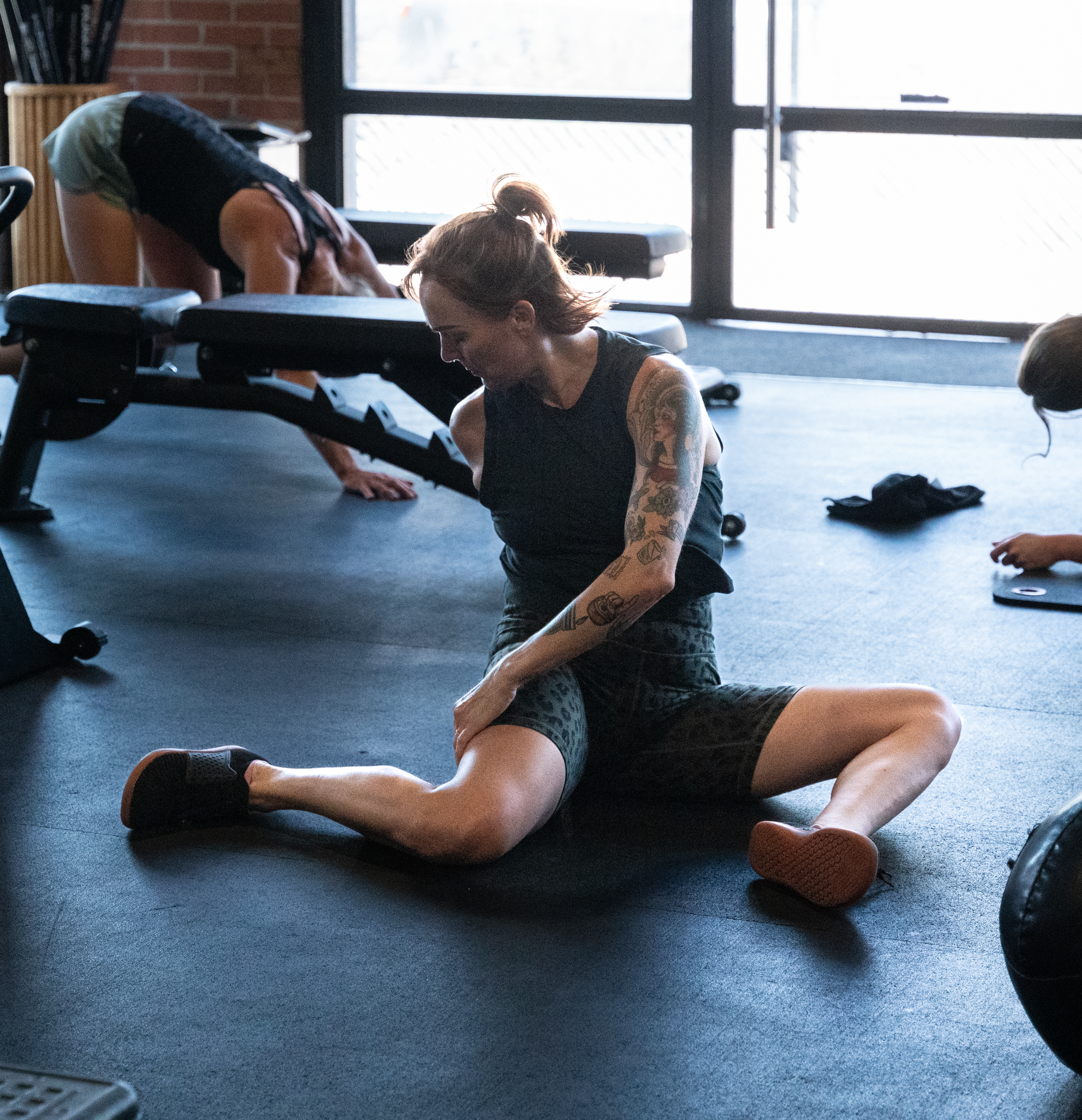 A woman with tattoos stretching on the gym floor while a person in the background is doing push-ups nearby.