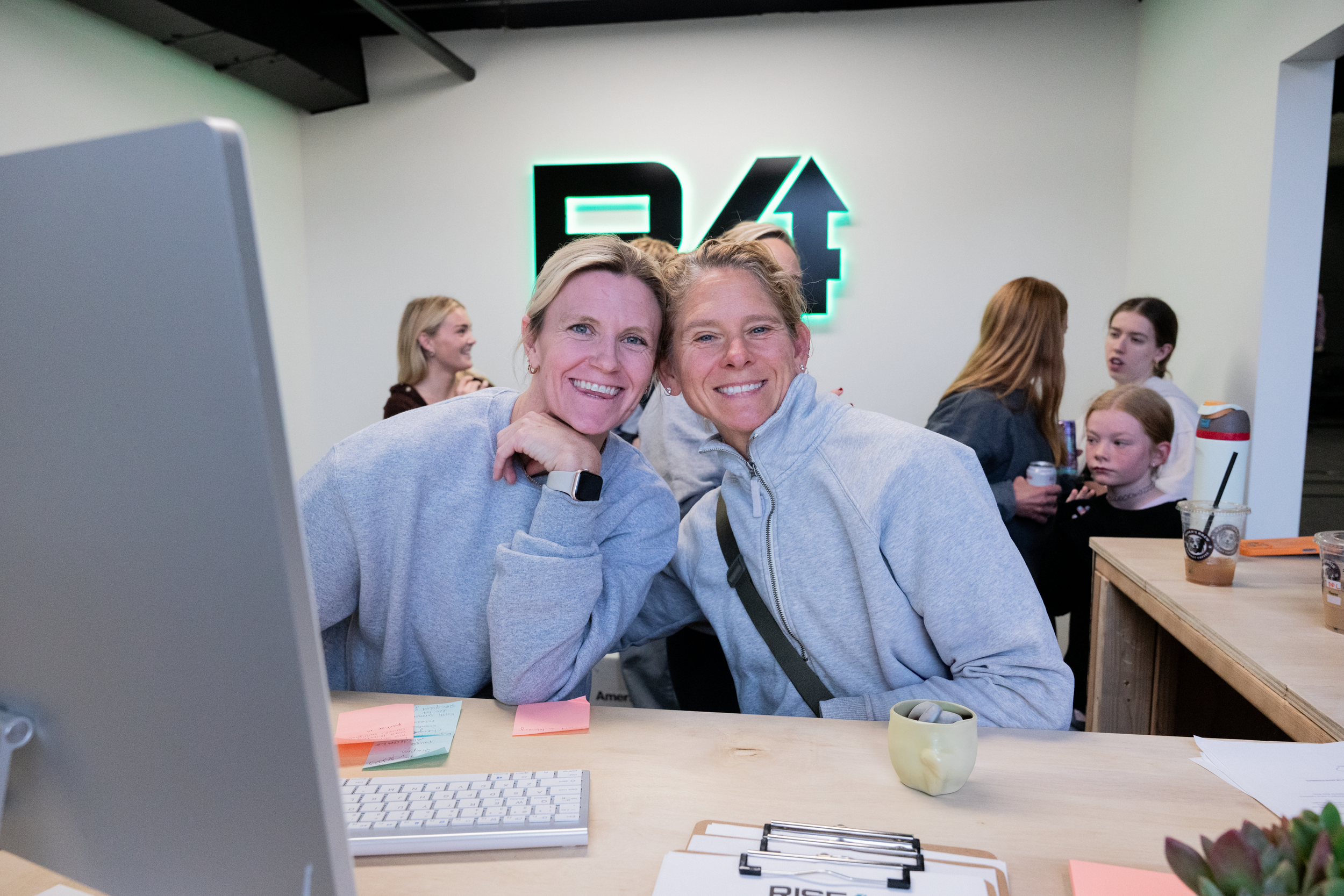 Two women smiling, sitting at a desk in an office, with a computer and notes in front. There are other people in the background, some talking and drinking beverages, with a neon sign on the wall behind them.