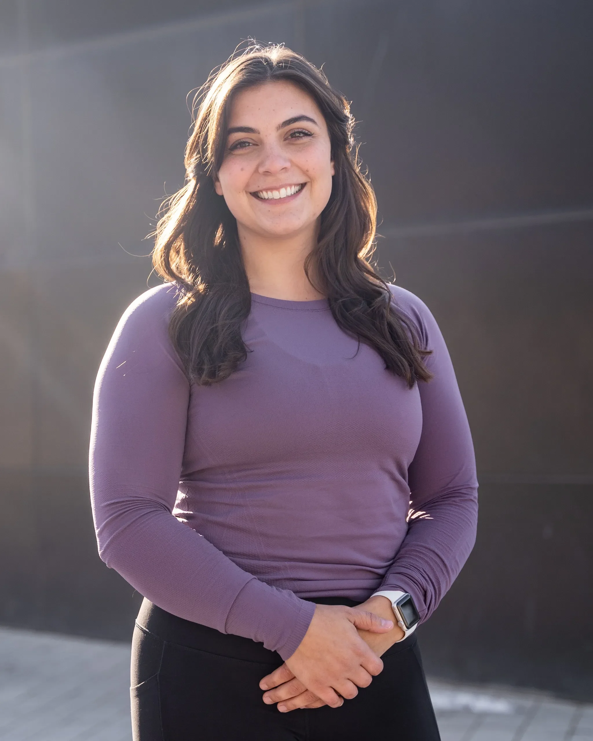 A woman with dark brown hair smiling outdoors, wearing a purple long-sleeve shirt and black pants, with a smartwatch on her wrist.
