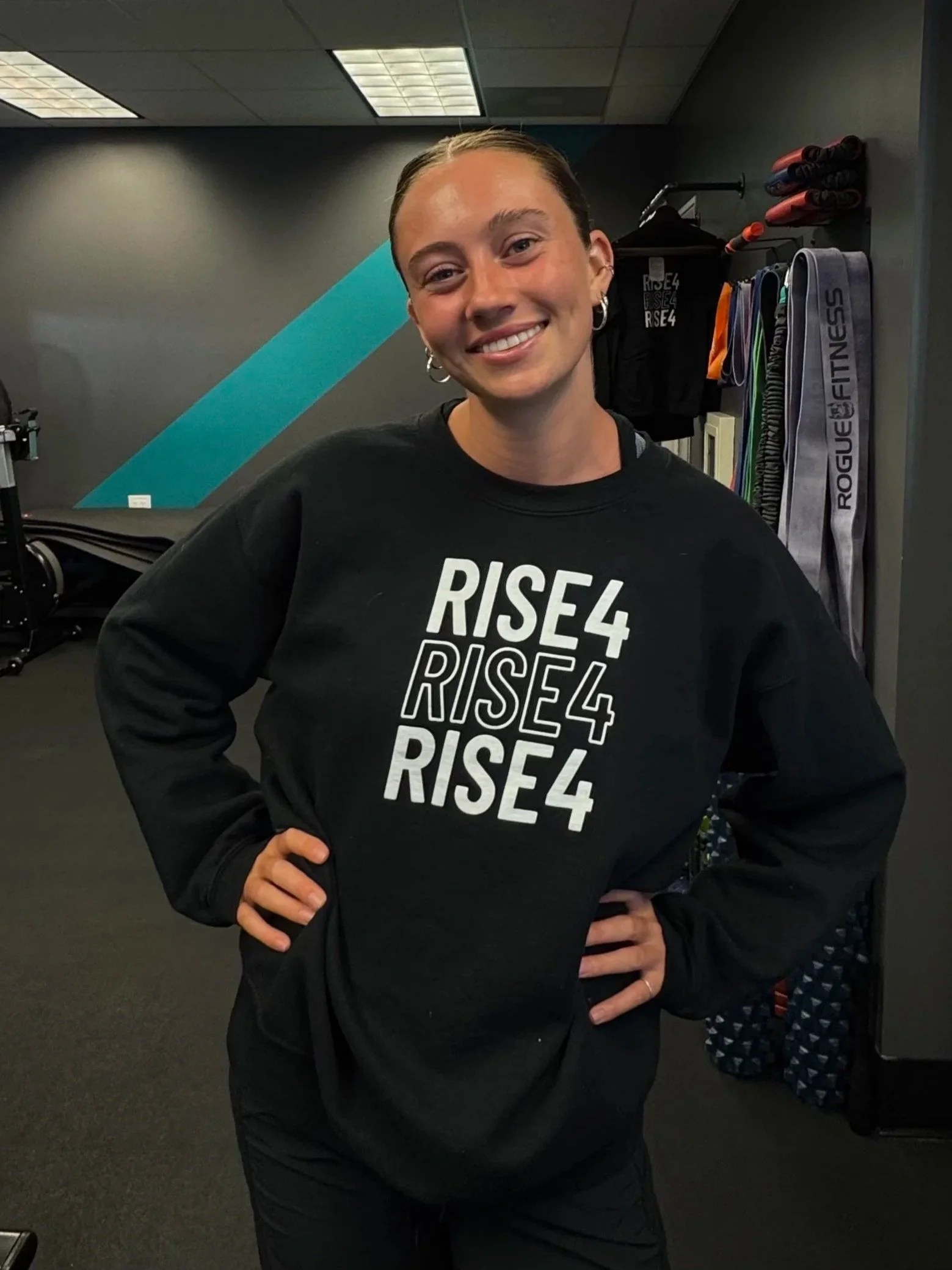Smiling woman with short hair, wearing a black sweatshirt with 'RISE4' written on it, standing in a gym.