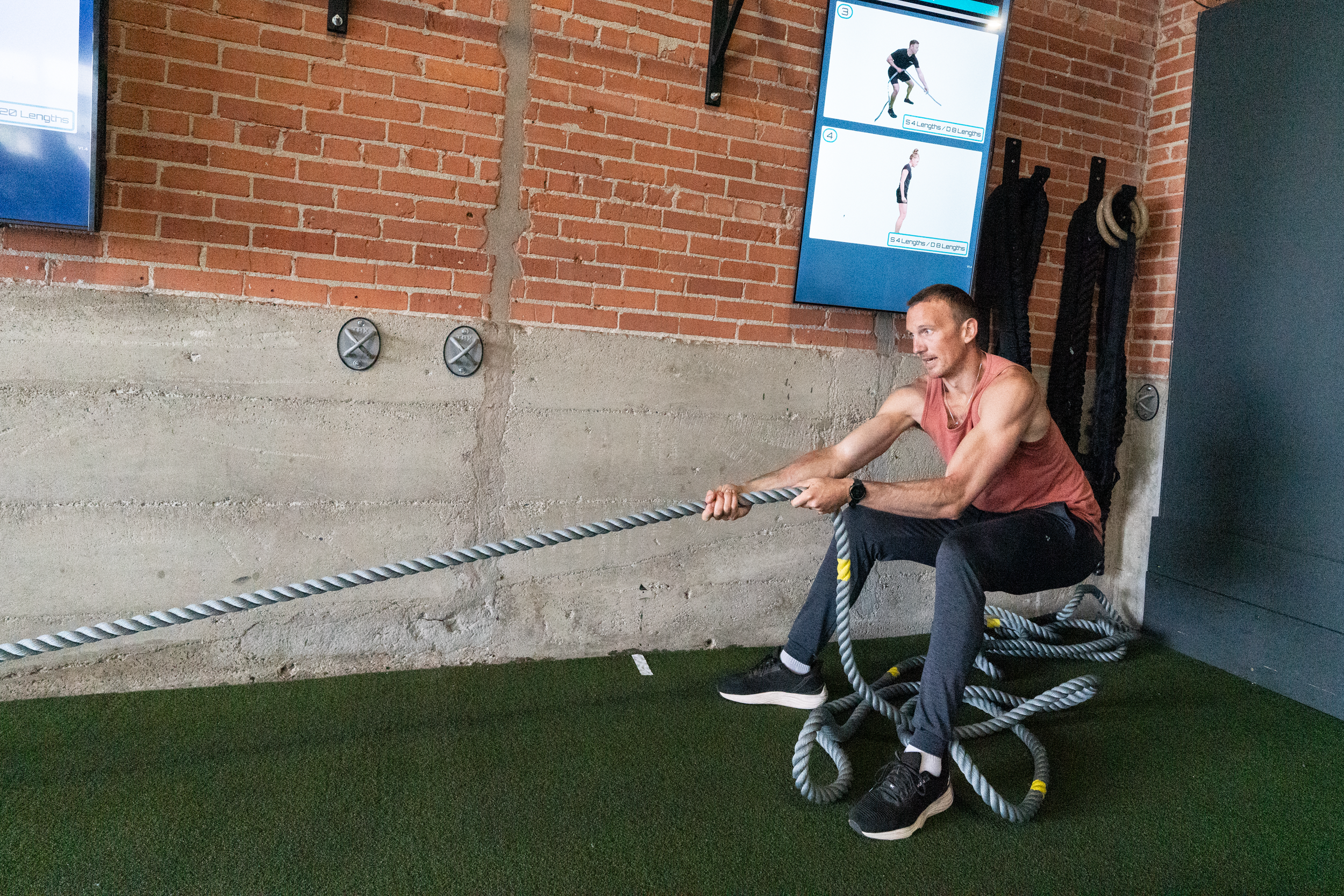 A man in a red tank top and black pants is sitting on the floor, pulling on a thick gray battle rope during a workout session at a gym. There are two large digital screens on the wall behind him displaying workout instructions with images of a woman and a man demonstrating exercises involving measuring lengths. The gym has a brick and concrete wall and is equipped with ropes hanging on the wall.