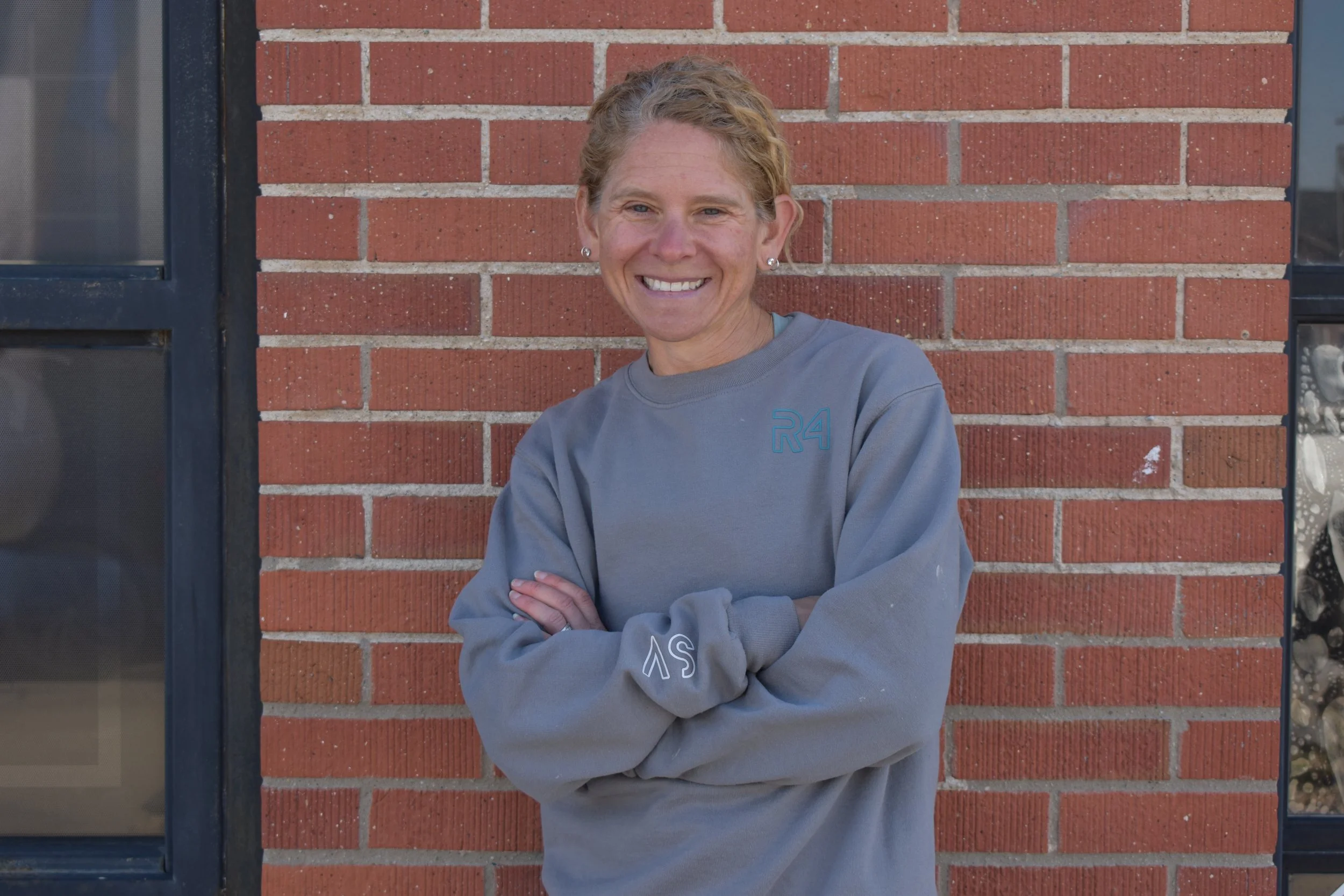 Woman with short curly hair smiling, standing with arms crossed in front of a brick wall, wearing a gray sweatshirt.