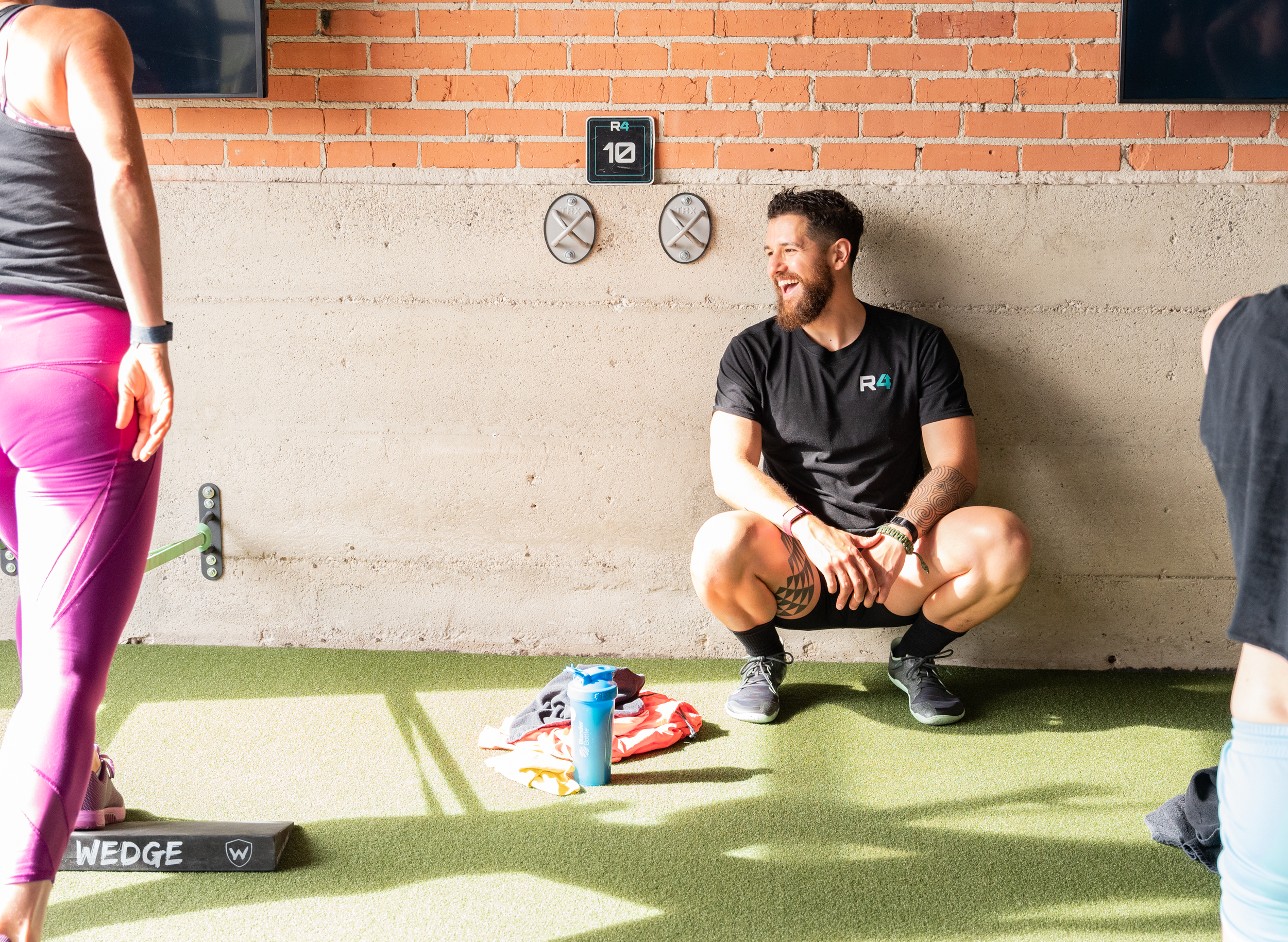 A man sitting against a brick wall in a gym, smiling, wearing a black T-shirt with the R4 logo, black shorts, and black sneakers. Two women are partially visible, one on each side, in workout clothing. The woman on the left is wearing pink leggings, and the woman on the right is wearing black shorts. On the ground in front of the man, there's a sports water bottle, a pile of workout clothes, and a balance board labeled 'WEDGE'.