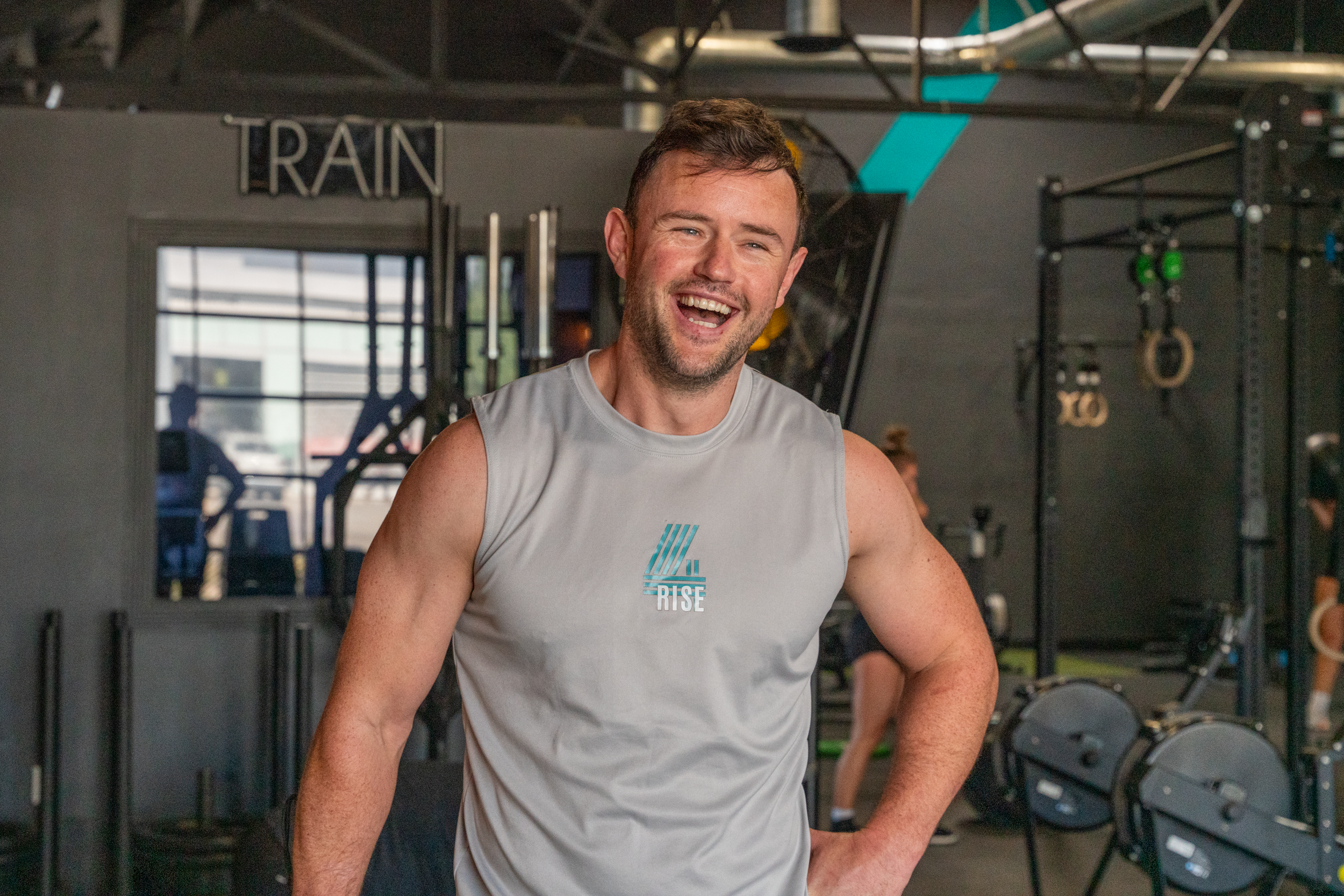 A smiling man in a gray sleeveless shirt inside a gym. The shirt has the words 'RISE' and a logo. The gym has workout equipment and a sign that says 'TRAIN' in the background.