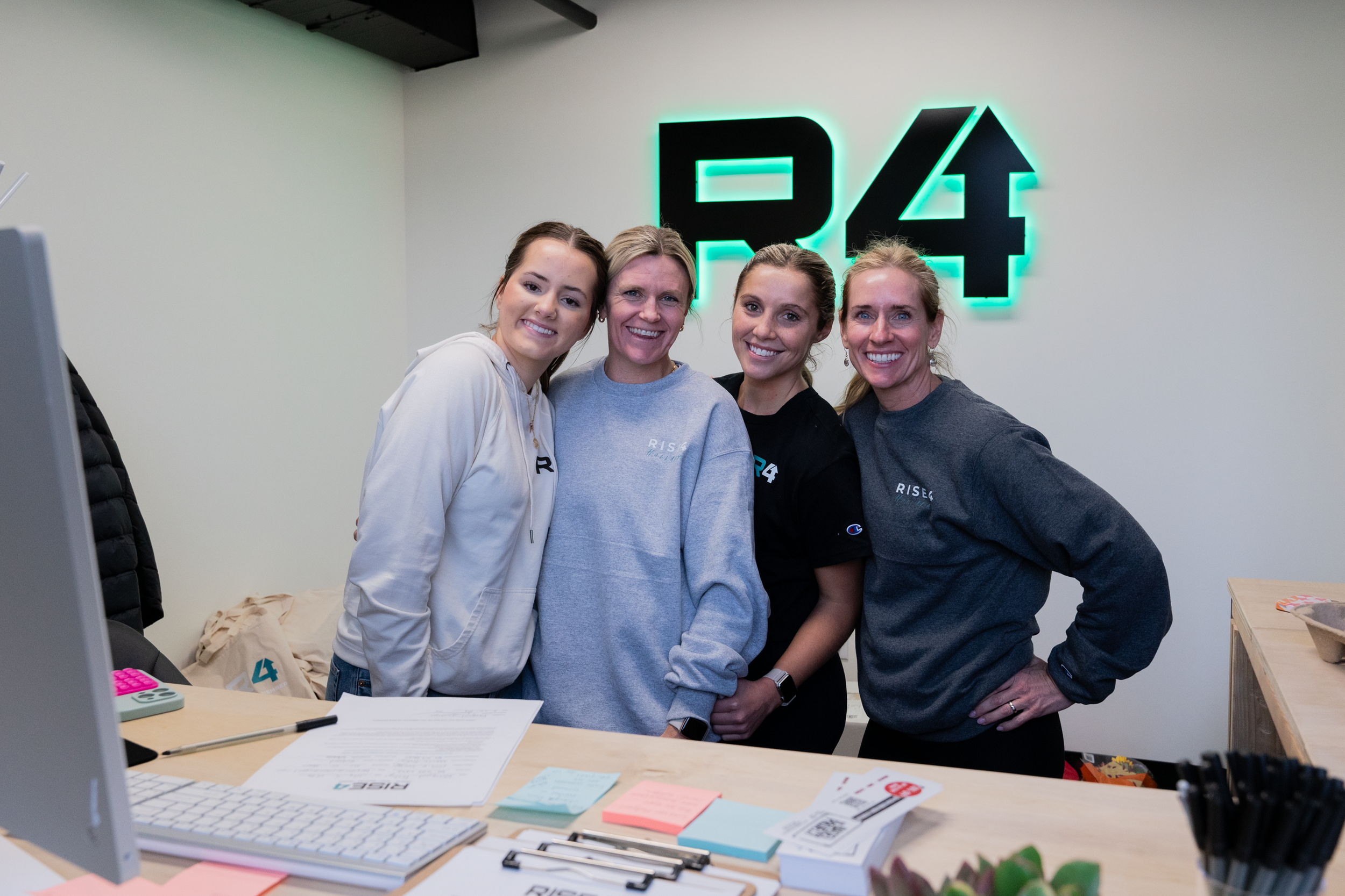 Four women standing together in an office, smiling, with a RISE4 sign on the wall behind them.