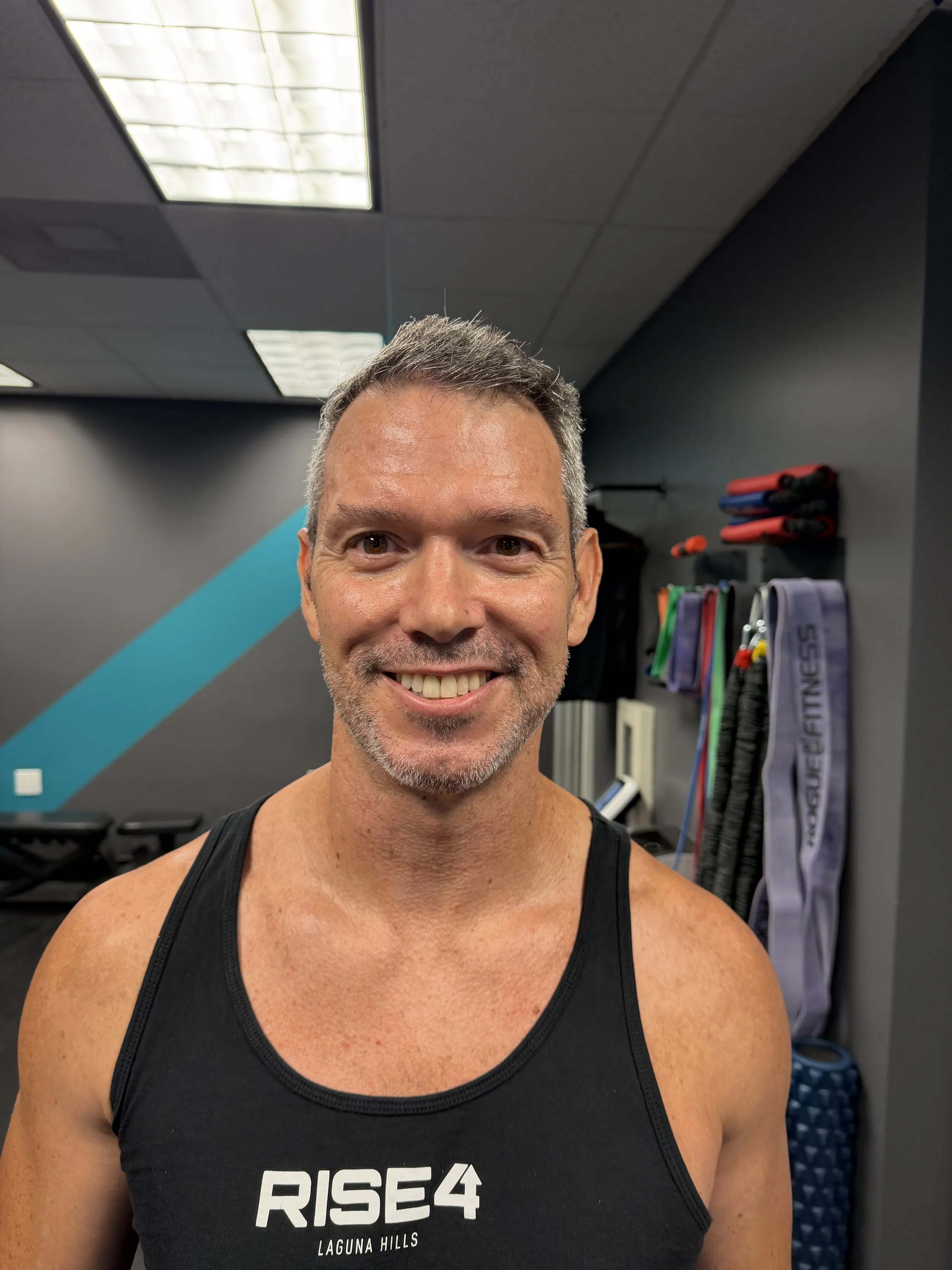 A smiling man in a black tank top with white text, standing in a gym with workout equipment and a dark wall in the background.