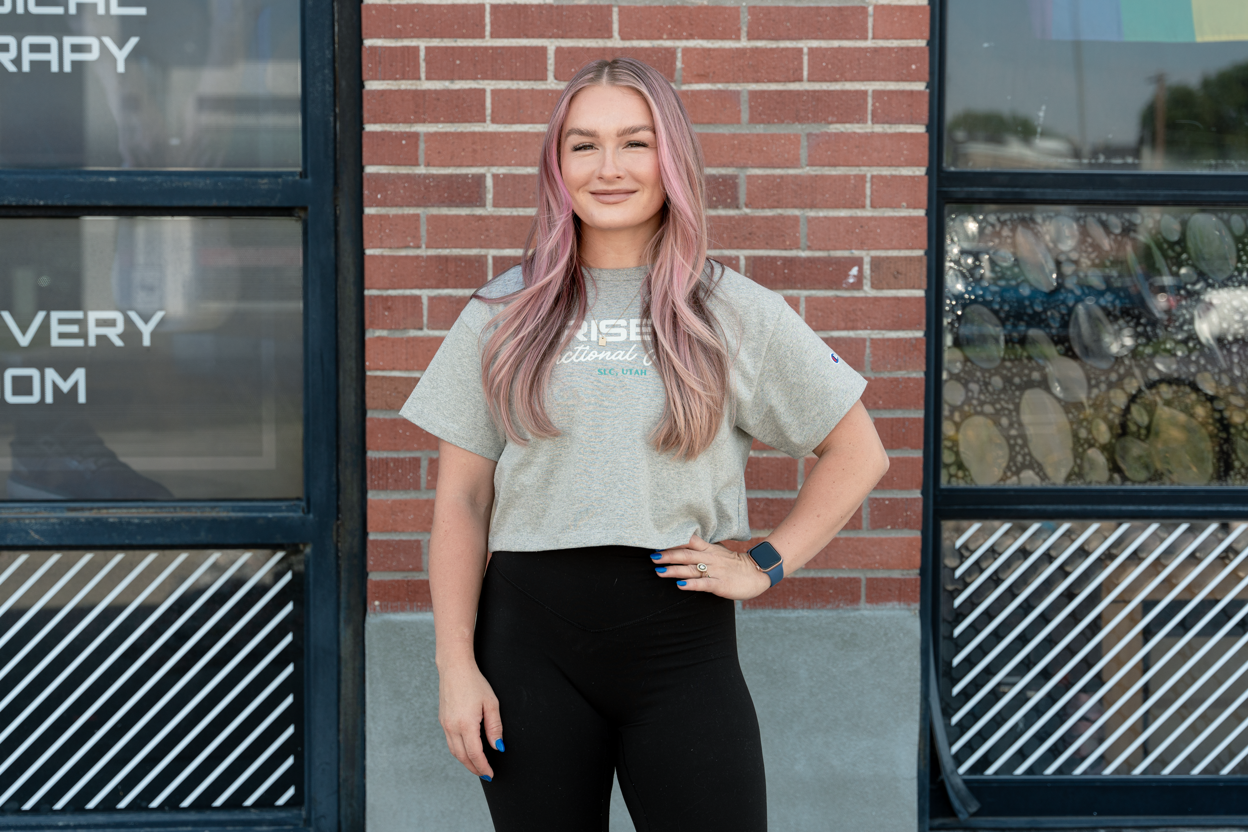 A young woman with pink hair standing outside against a brick wall, smiling, wearing a gray t-shirt and black pants.
