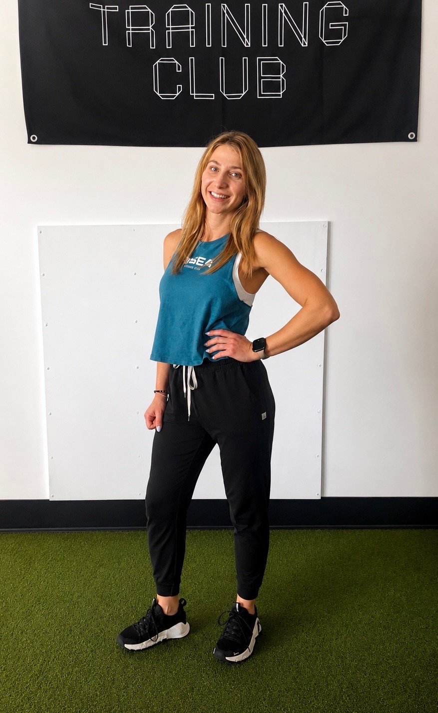 A smiling woman in athletic clothing standing inside a gym with a black "Training Club" banner on the wall behind her.