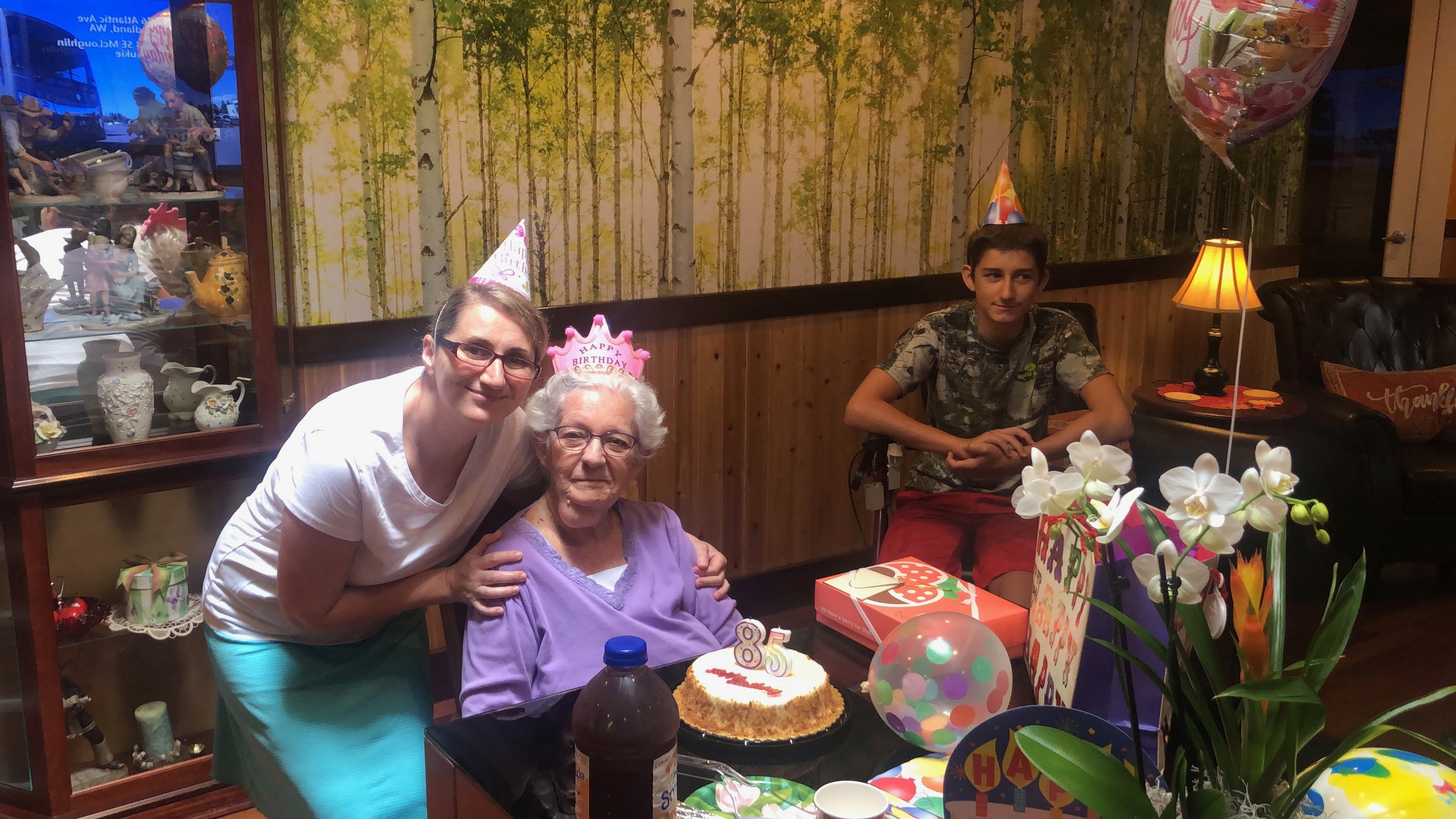 Elderly woman at table with birthday cake, surrounded by friends or family celebrating her 85th birthday. Party decorations, balloons, and flowers are present.
