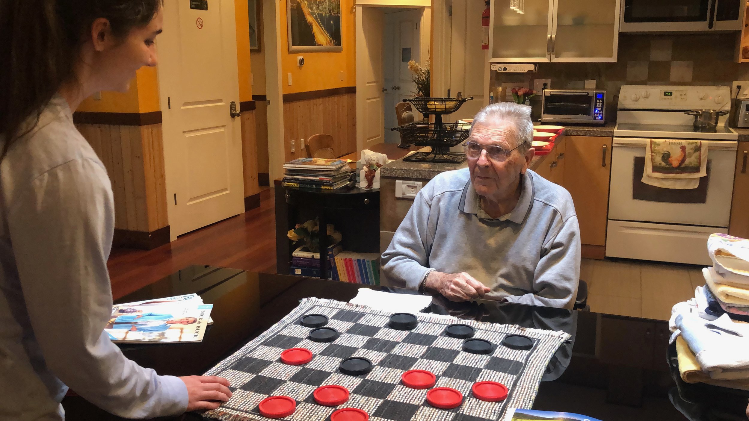 An elderly man playing checkers with a young woman in a kitchen setting.