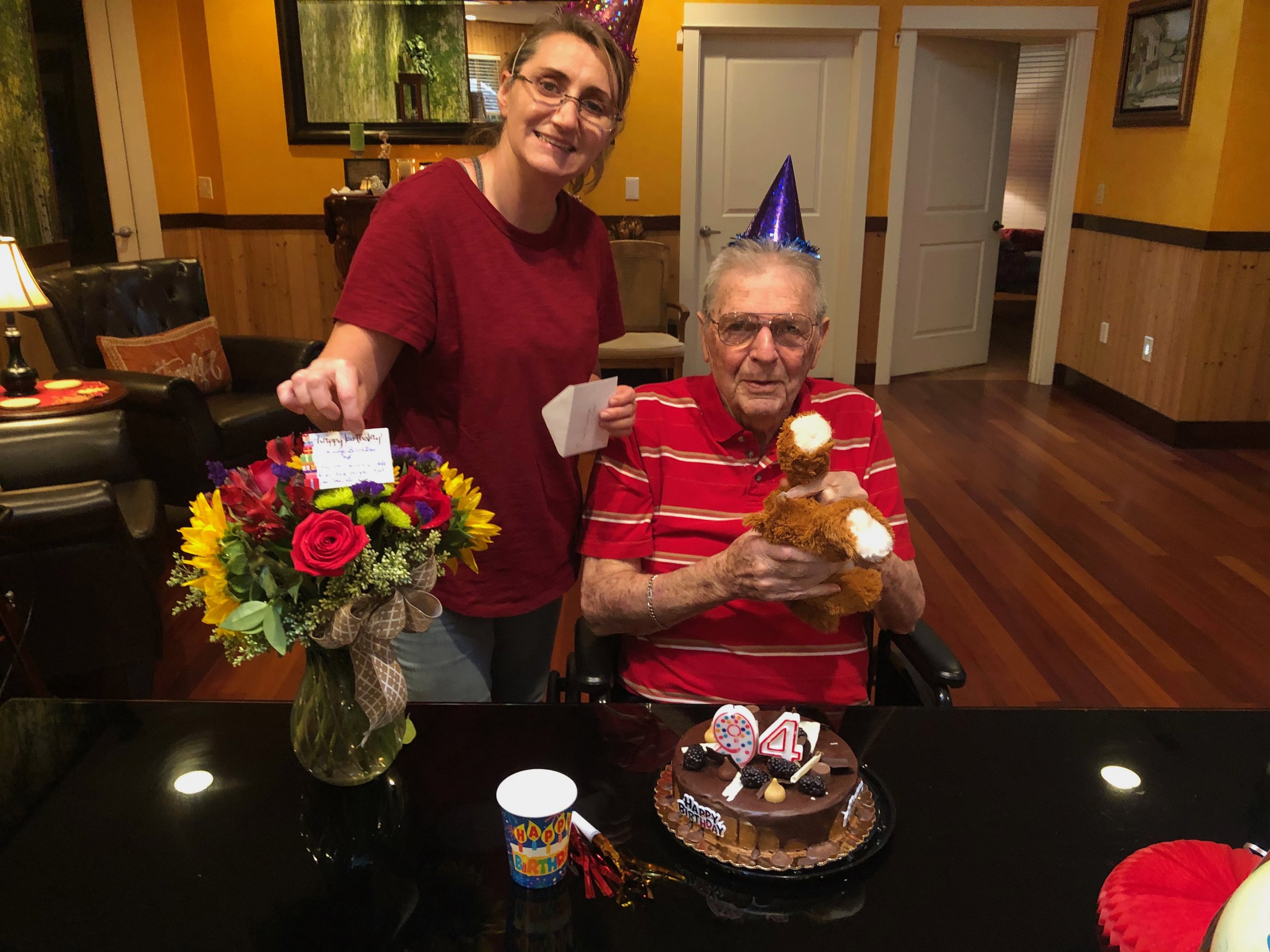 Birthday celebration with a man in a striped shirt and party hat holding a teddy bear. A woman in a red shirt and party hat stands next to him holding a bouquet of flowers with a card. A chocolate cake with "94" candles is on the table.