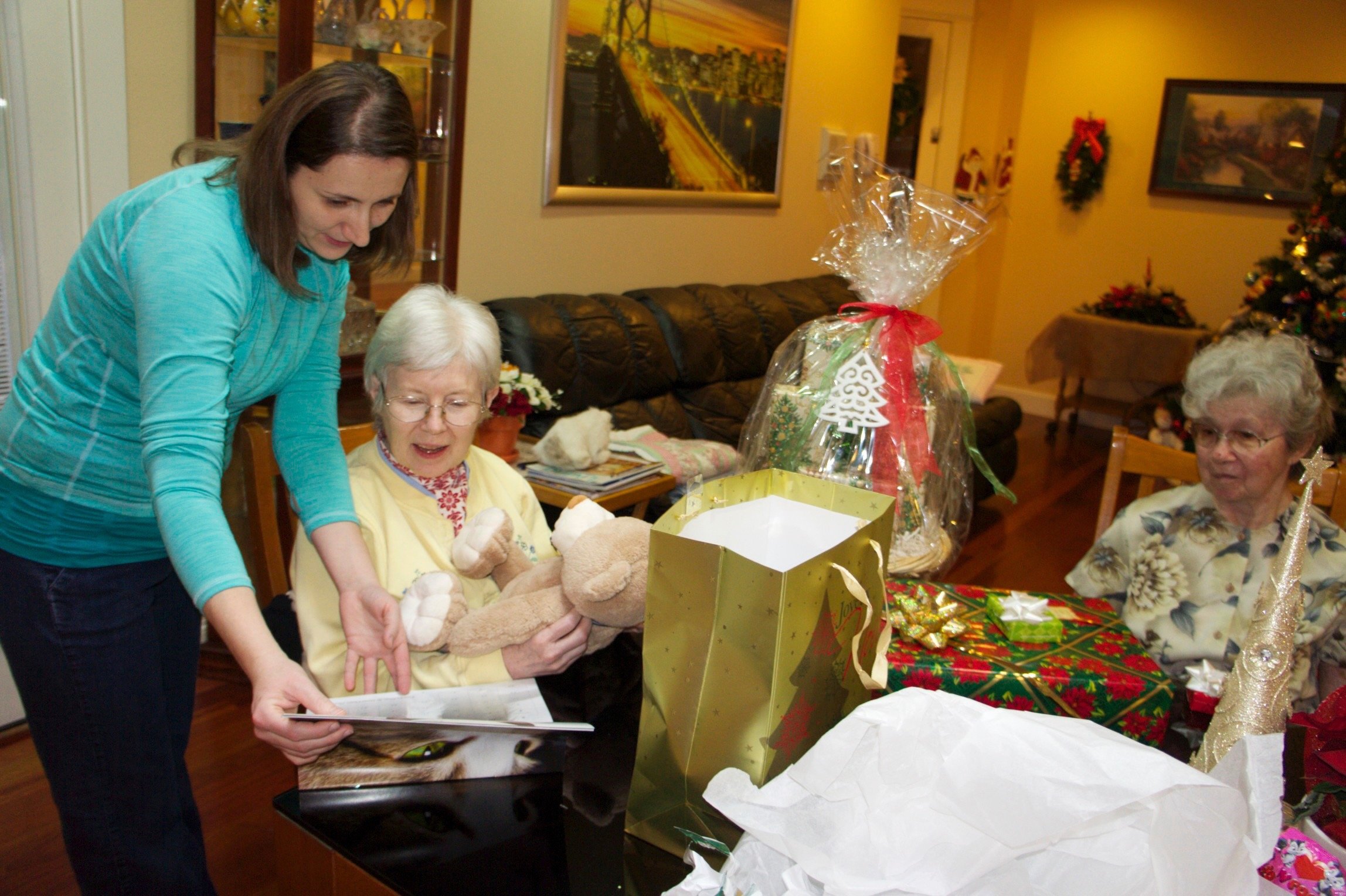 Three women in a living room, two older women seated at a table with gifts, including a stuffed animal, gift bags, and wrapped presents. The room is decorated for Christmas with wreaths and a tree in the background. One woman is showing an item to an