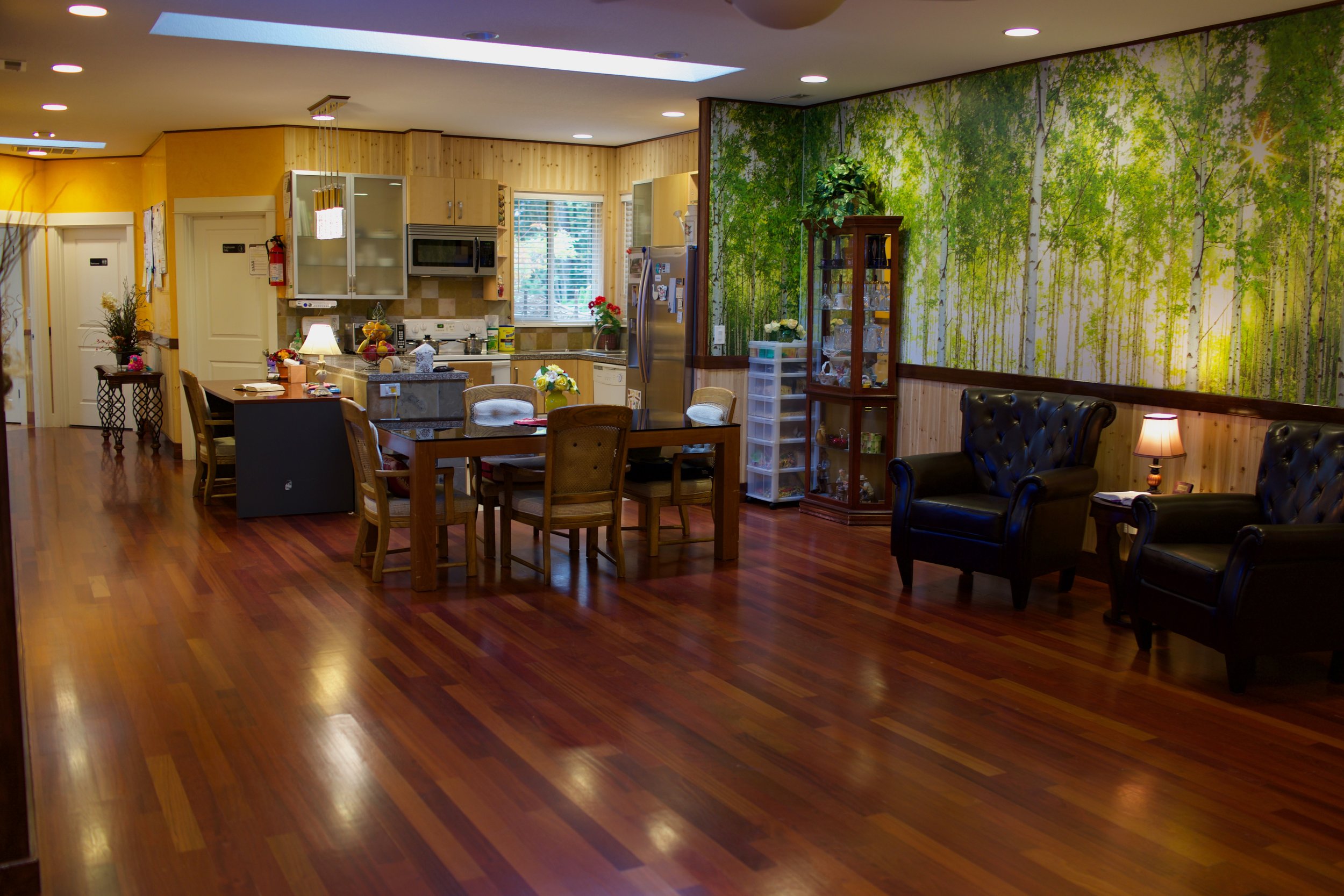 Open kitchen and living area with wooden flooring, dining table with chairs, and kitchen appliances. Wall mural of a forest scene and black leather armchairs near a small lamp and glass cabinet.