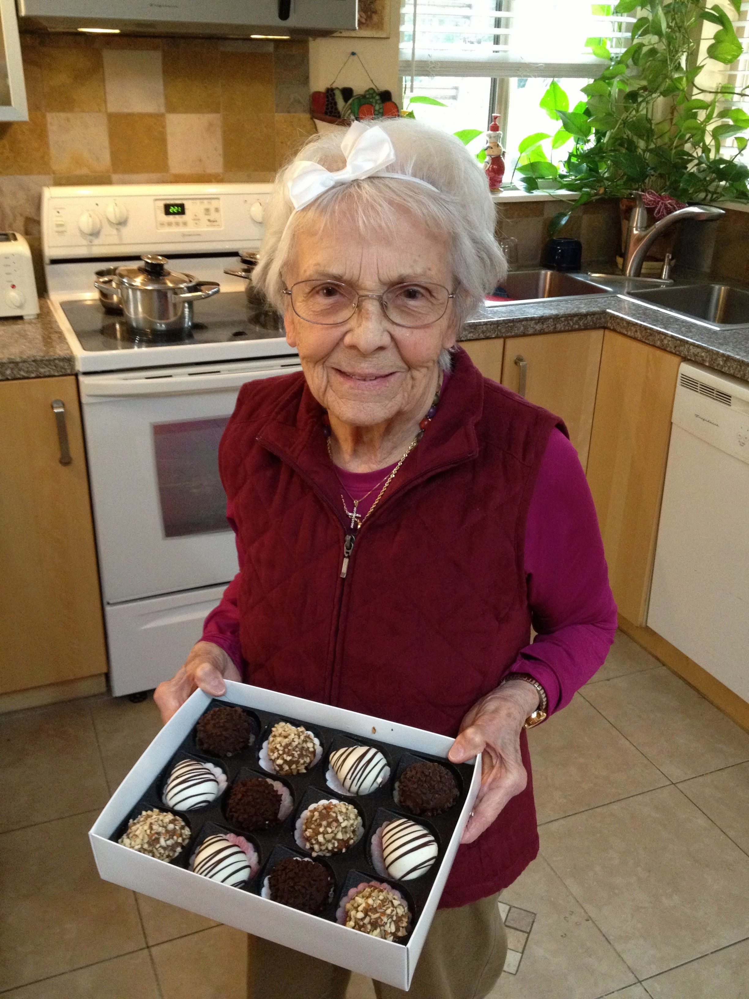 Elderly woman holding a box of assorted chocolates in a kitchen with plants and a stove in the background.
