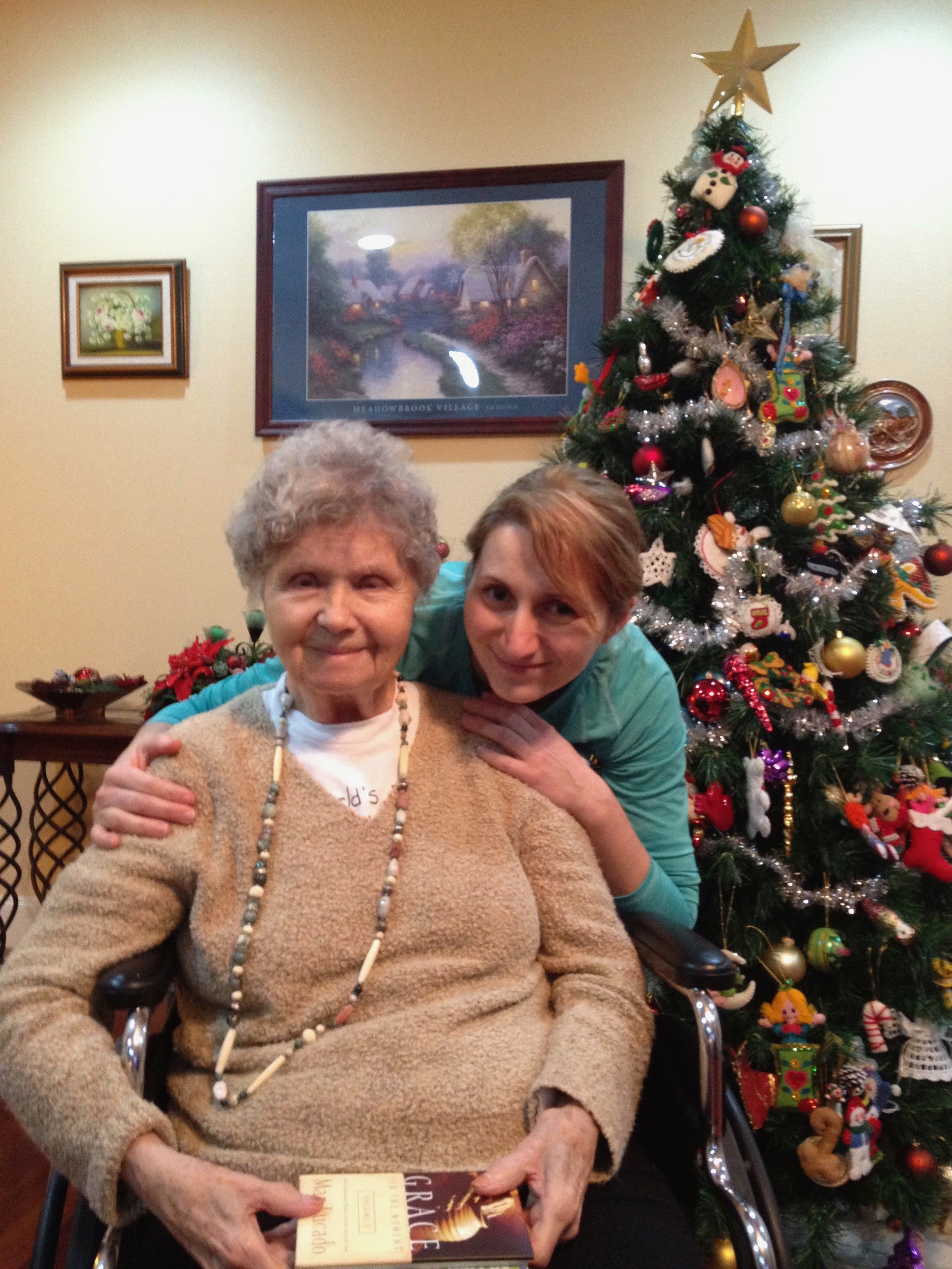 Two people smiling in front of a decorated Christmas tree. The older person is seated, holding a book, while the younger one is standing beside them with an arm around their shoulder. Framed paintings are visible in the background.
