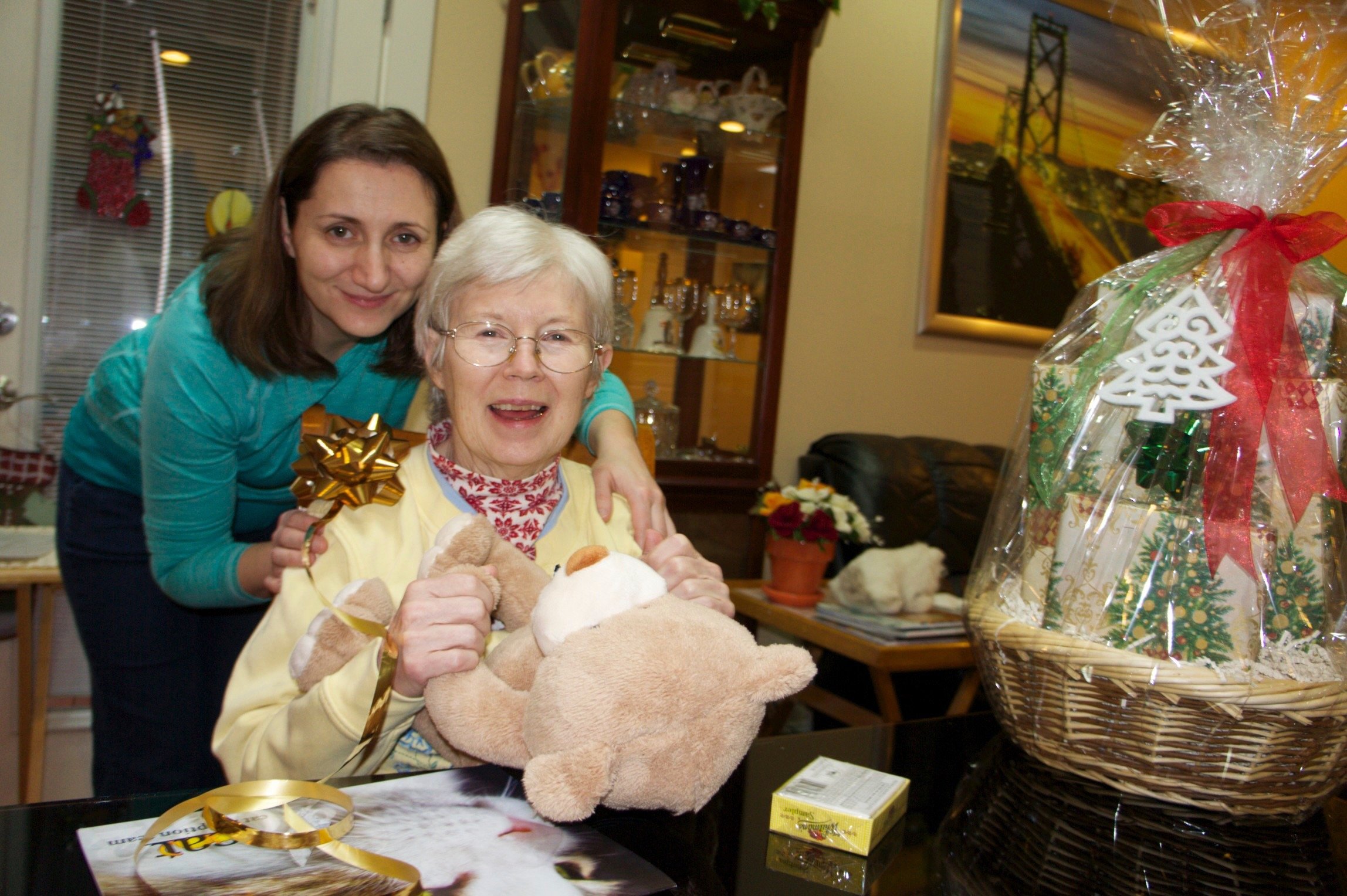 Two women sitting at a table, one holding a plush bear and a gold bow. A large gift basket wrapped in plastic with red and green decorations sits on the table. A display cabinet and framed picture are in the background.