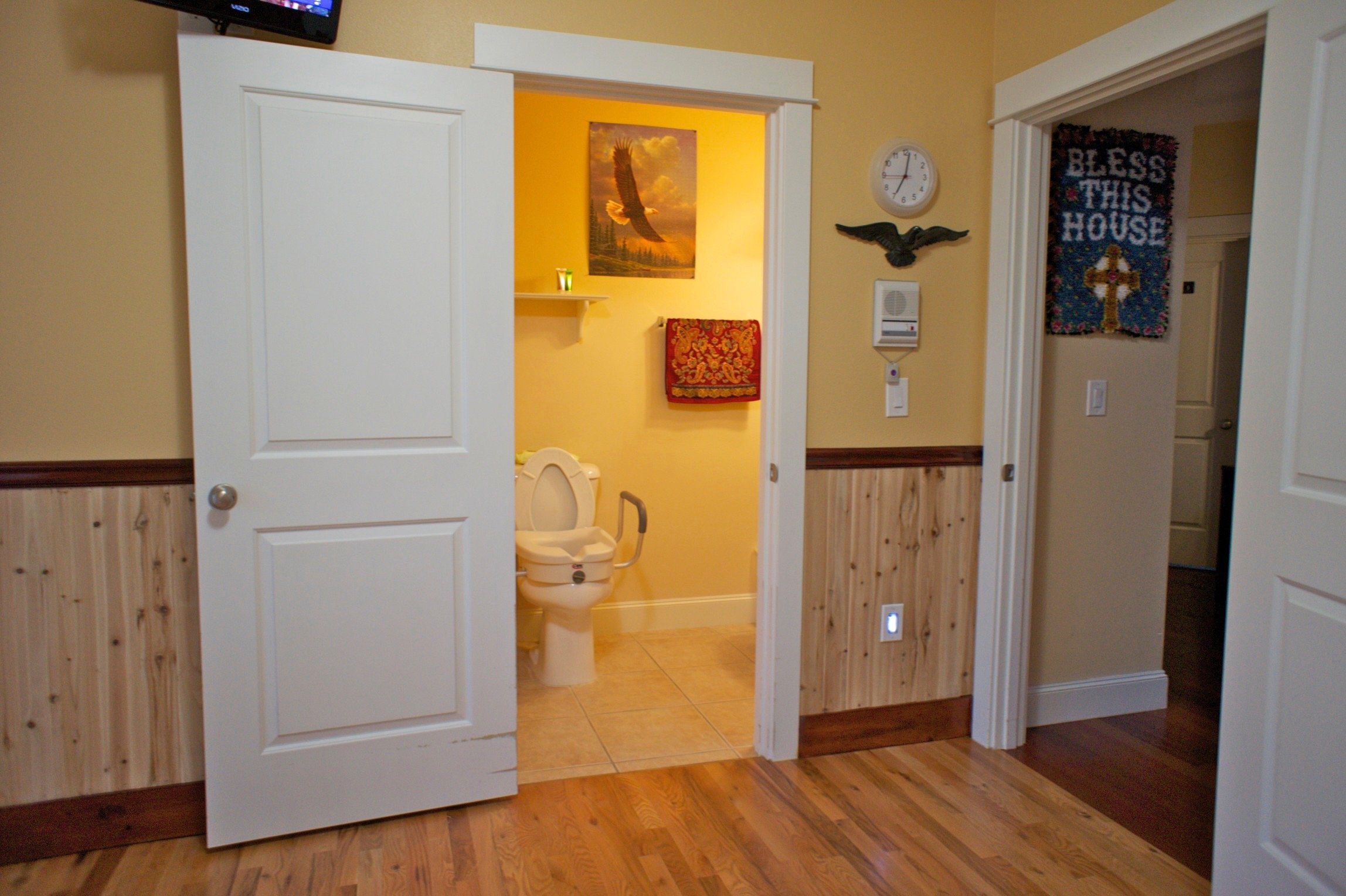 A bathroom with a white toilet equipped with safety handles. The room has yellow walls and wooden flooring. A framed picture of an eagle is on the wall above a shelf with a candle. A red patterned towel hangs beneath the shelf. Outside, a wall clock,