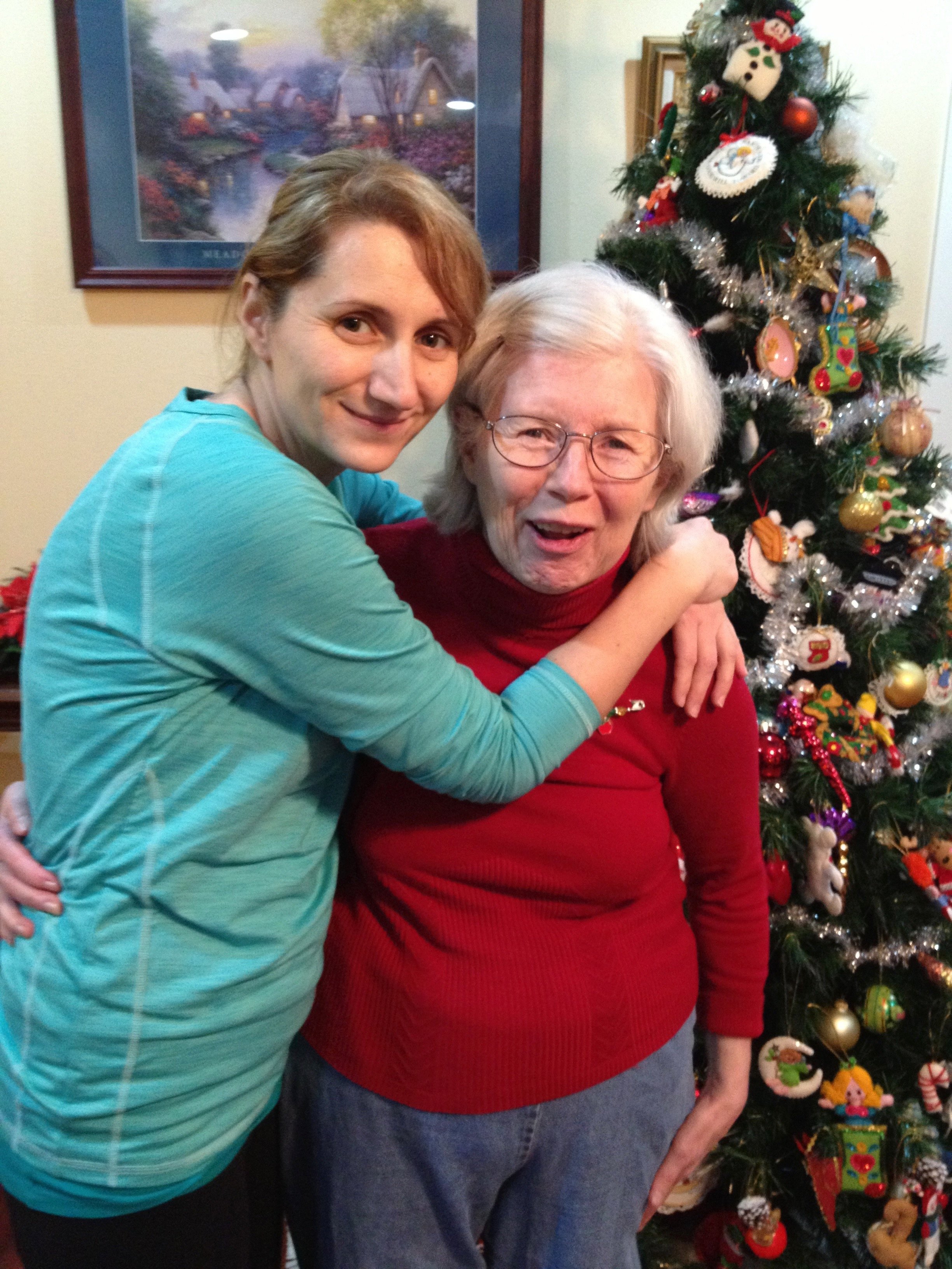 Two women posing happily in front of a decorated Christmas tree, one wearing a blue sweater and the other in a red sweater. The room is warmly decorated with a painting on the wall.
