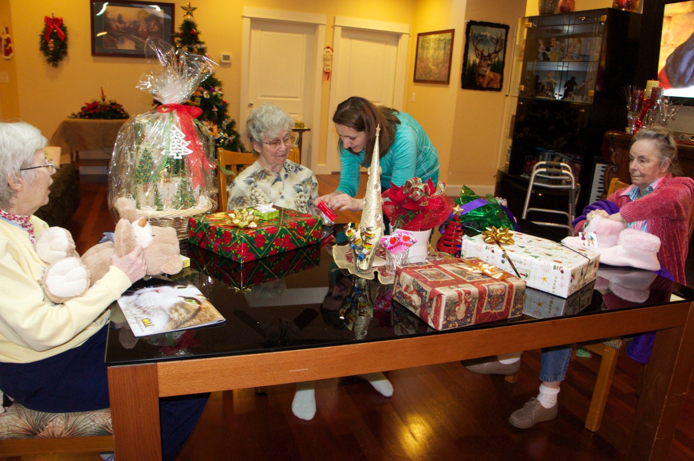 Group of elderly women and a caregiver sitting at a table with Christmas gifts, decorations, and a holiday setting in the background.