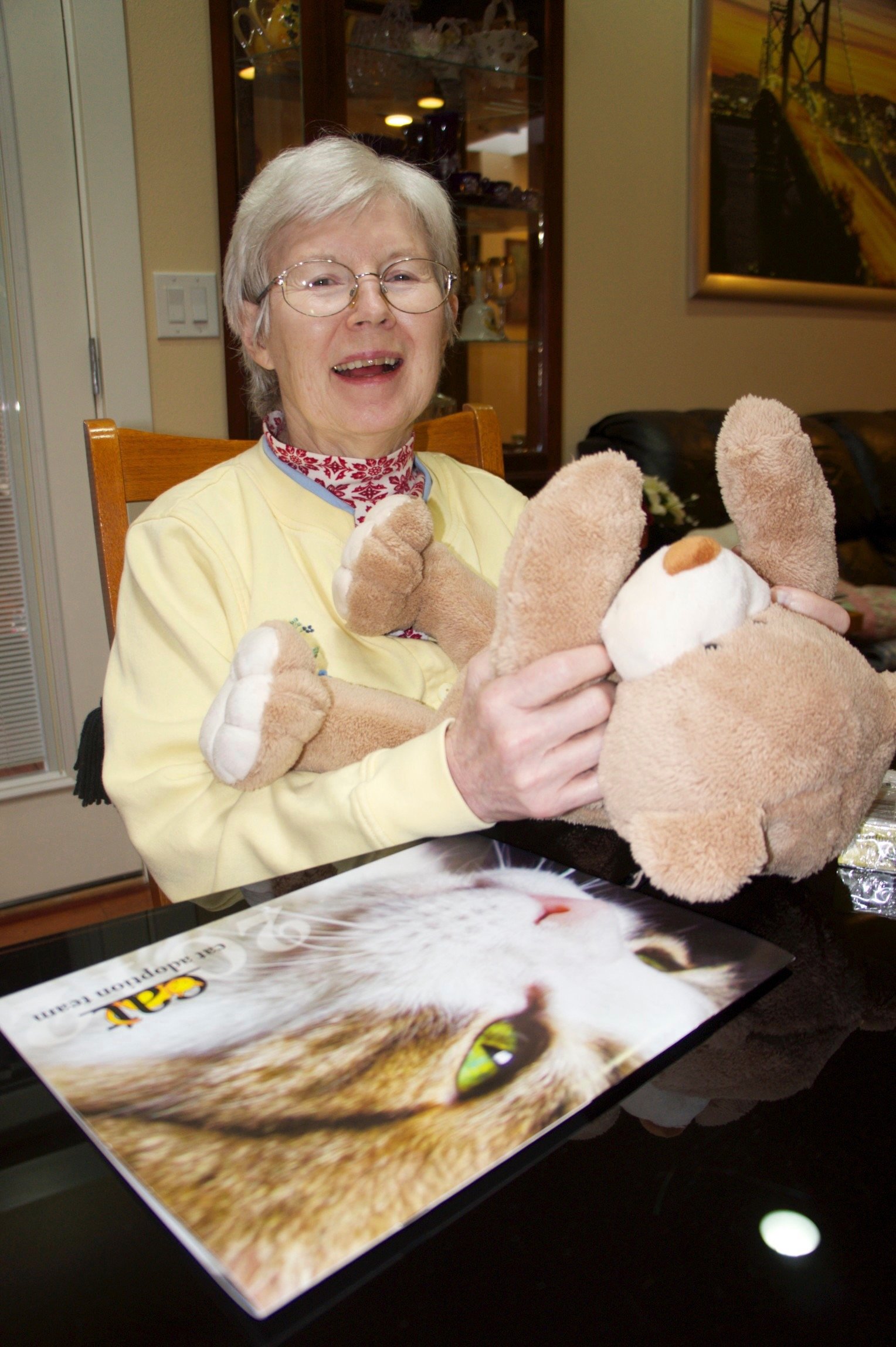 Elderly woman holding a teddy bear next to a large cat-themed magazine.
