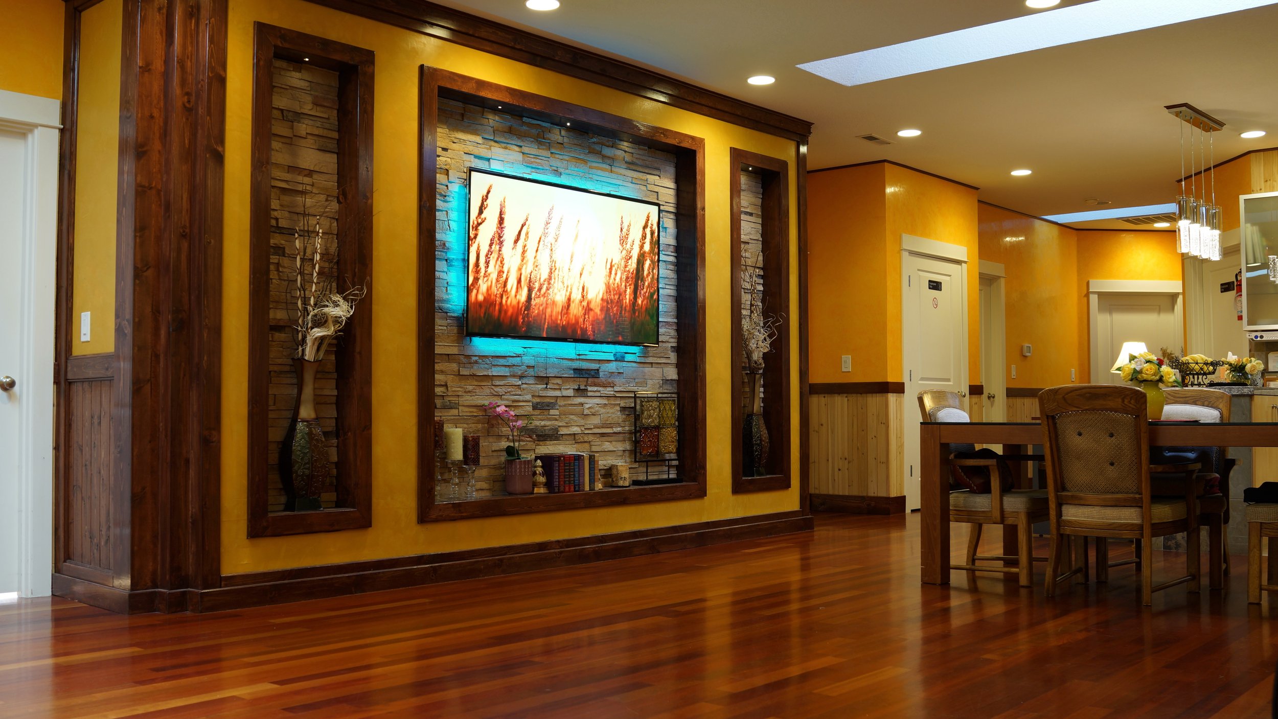 Interior of a room with hardwood floors, yellow walls, and a wooden dining table. A wall-mounted TV is displaying a landscape image. Decorative vases and small plants are arranged nearby. Recessed lighting and pendant lights illuminate the space.