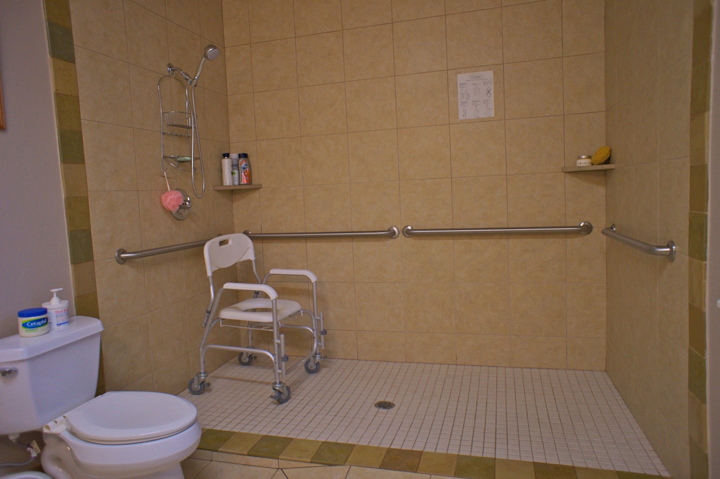 Accessible bathroom with a shower area, grab bars, and a shower chair; also visible are a toilet, shelves with toiletries, and a tiled floor.
