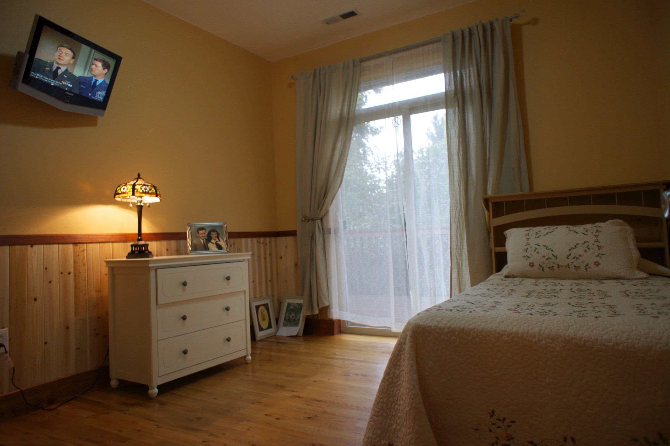 Bedroom with wooden floor, single bed with floral bedding, dresser with lamp and photos, wall-mounted TV, and large window with curtains.