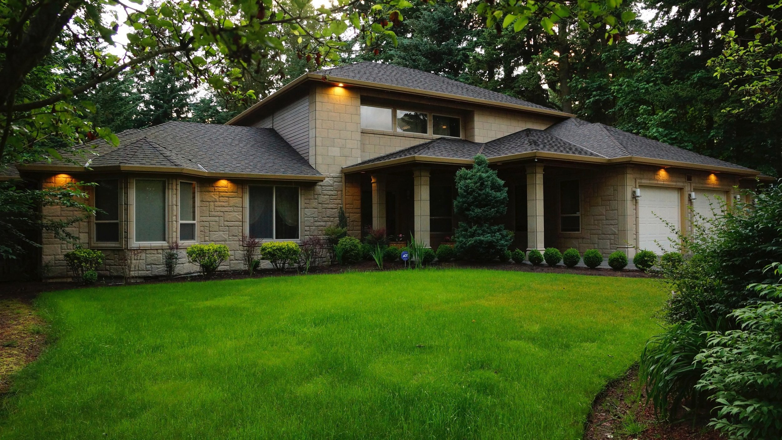 A modern two-story house with a stone exterior, landscaped lawn, and a neatly trimmed hedge in front. The house features a large garage and multiple windows, surrounded by tall trees.
